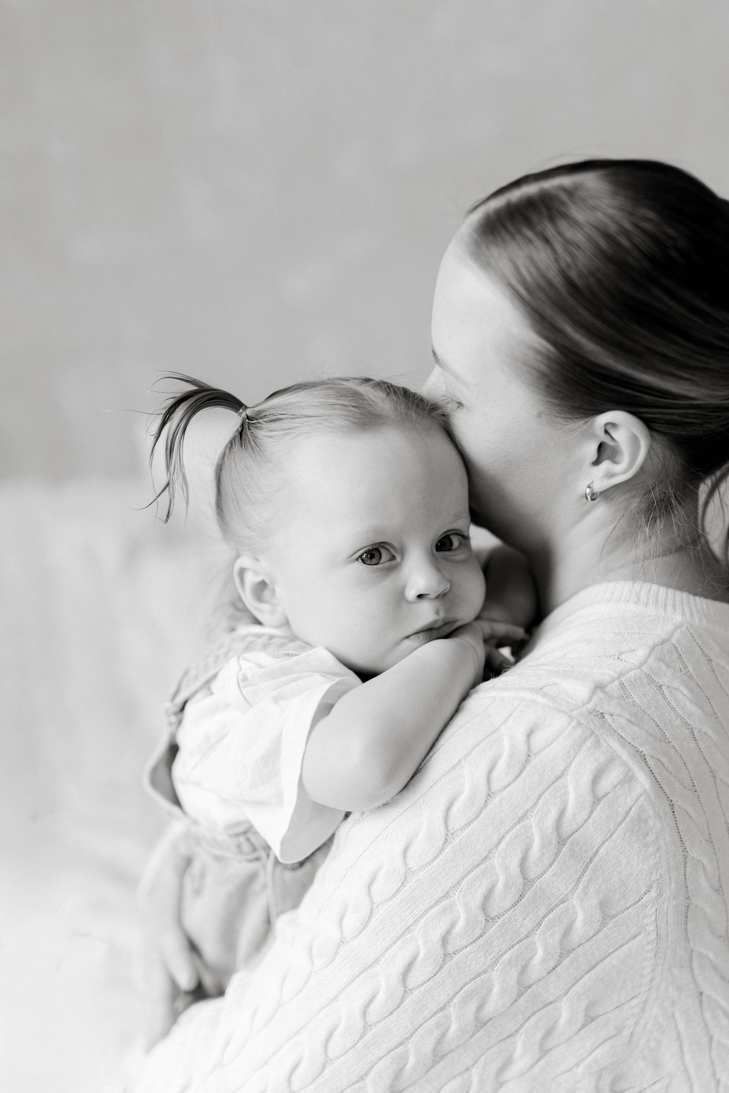 Black and white photo of a mother kissing her baby girl with pigtails, who looks at the camera.