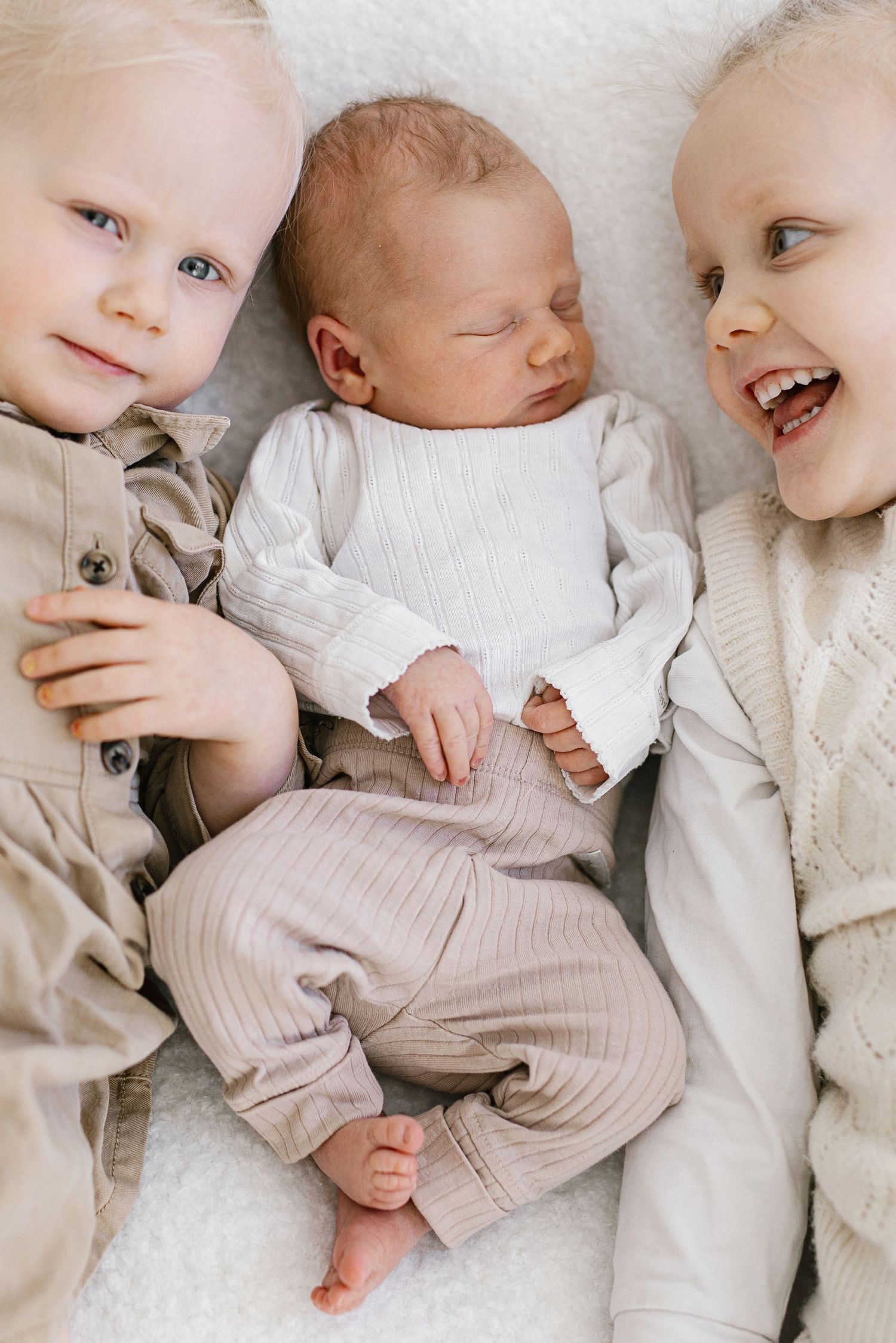 Two babies in neutral colored outfits lying together on white bedding.