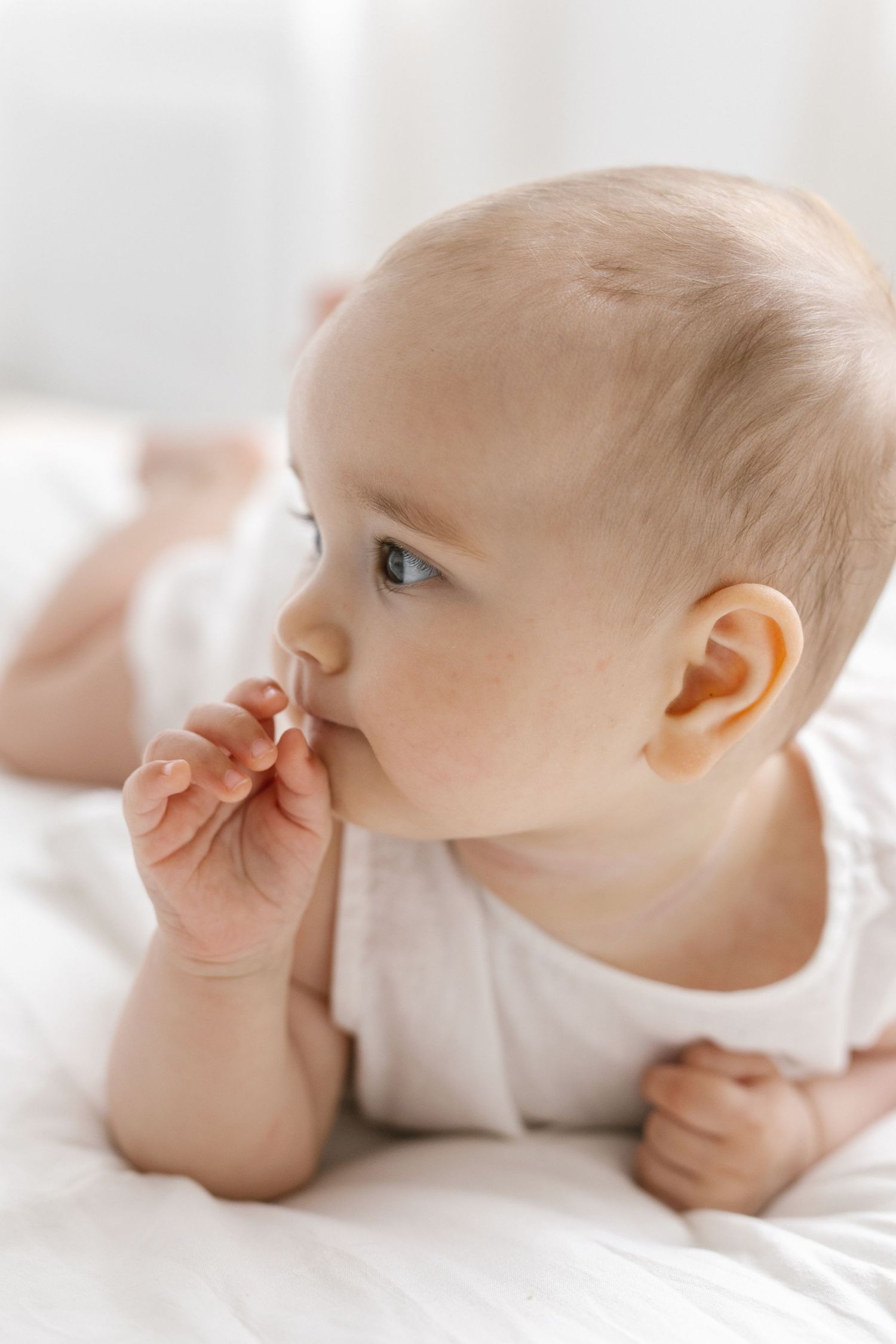 Baby in a white onesie lying on a soft white bed, chewing a tiny finger while gazing to the side in bright natural light.
