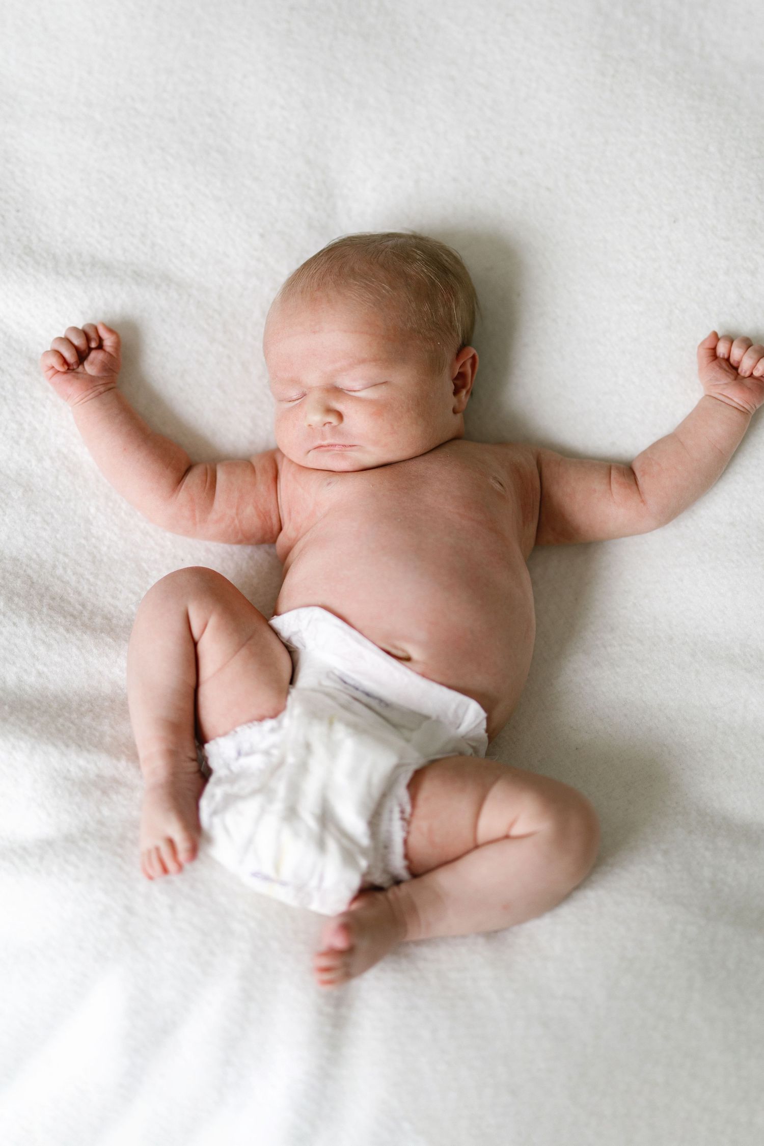 Newborn baby sleeping peacefully on white blanket wearing diaper with arms raised in startle reflex position.