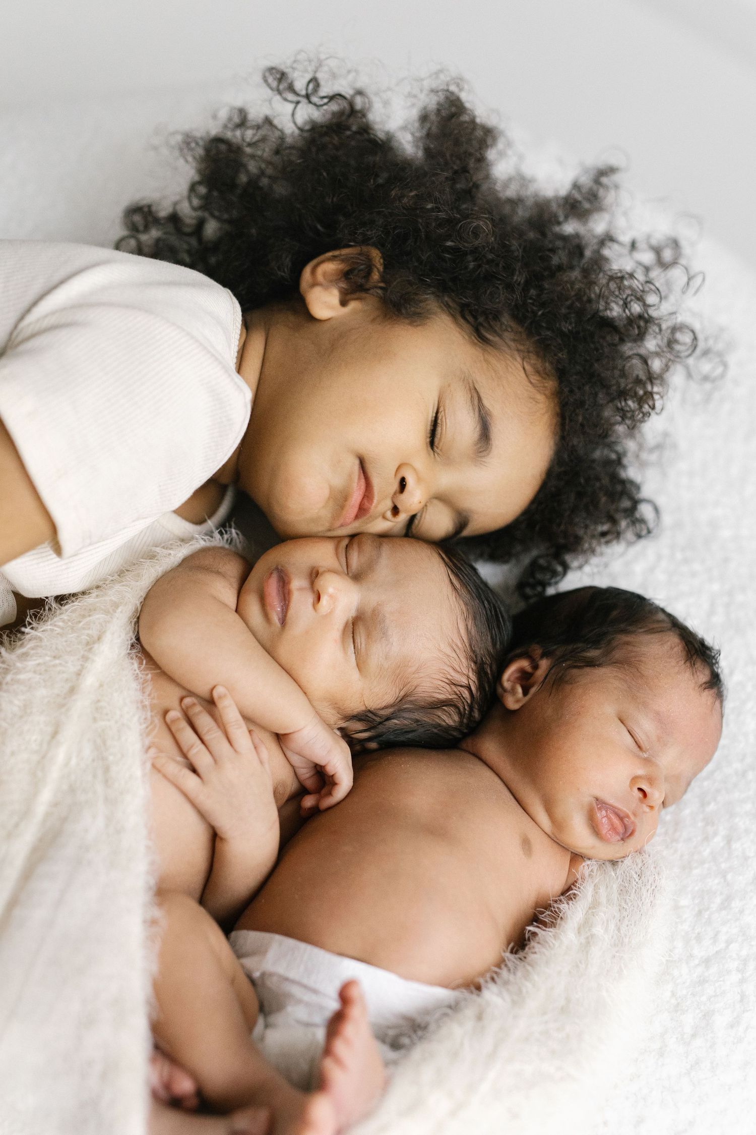 A young girl with curly hair gently kisses two sleeping newborn twins lying on a soft white blanket.