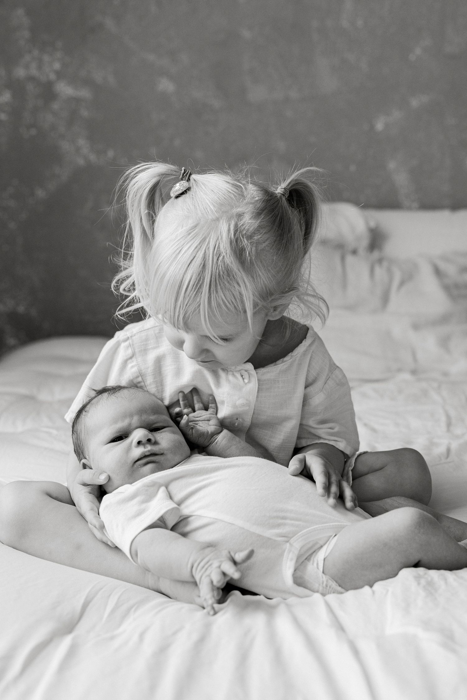 Black and white photo of a young girl with pigtails gently holding a newborn baby on a white bed.