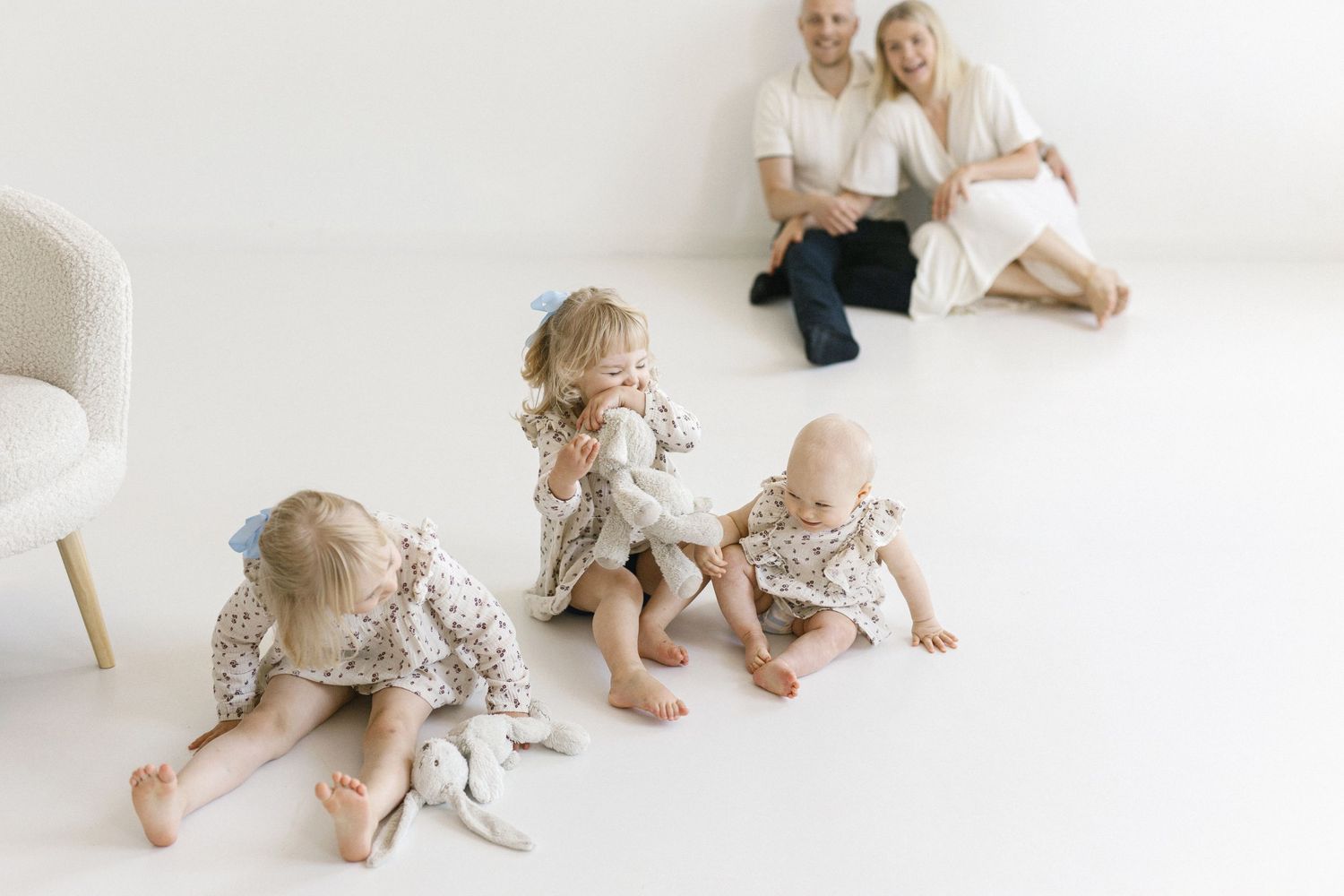 Three babies in floral outfits play on a white floor while their smiling parents sit together in the background.