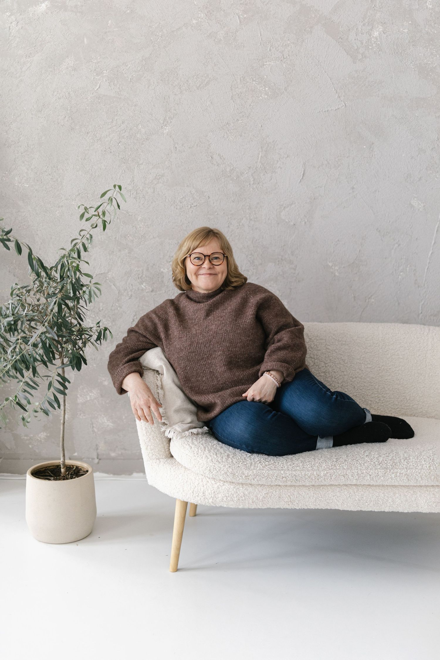 A smiling middle-aged woman in a brown sweater and jeans sits relaxed on a white sofa beside a potted plant.