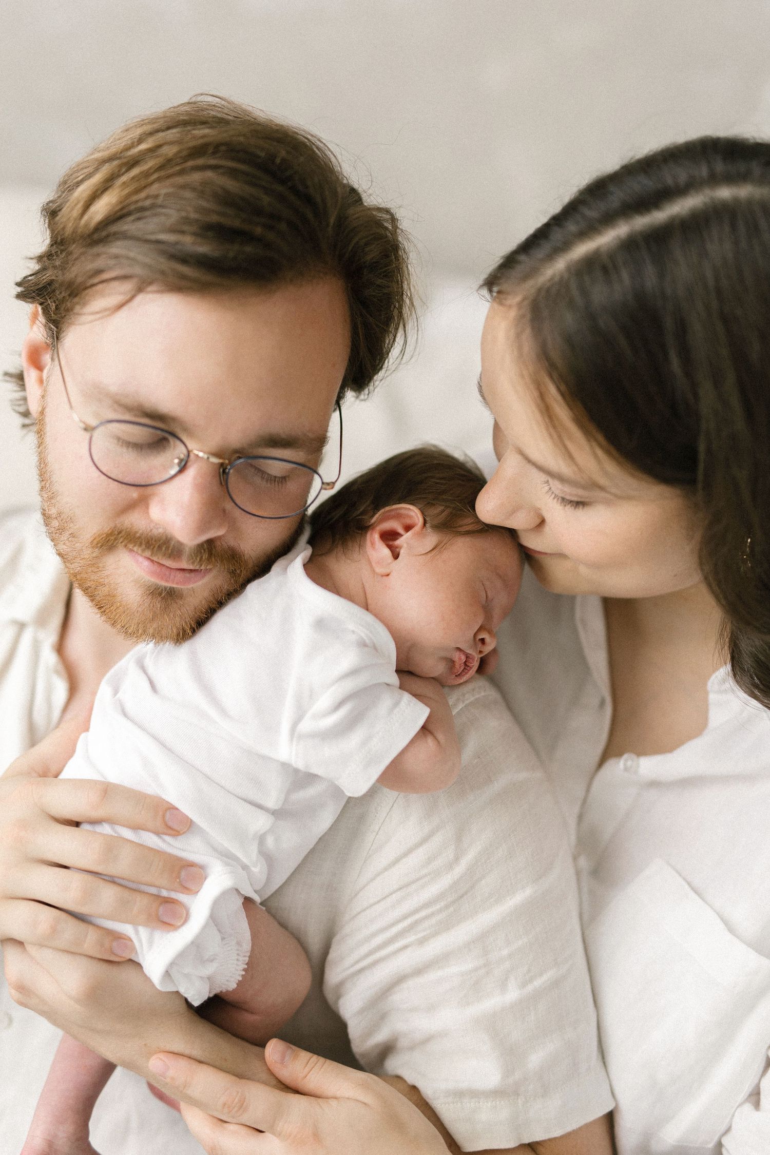 A couple in white shirts tenderly holds and kisses their newborn baby during an intimate family portrait session.