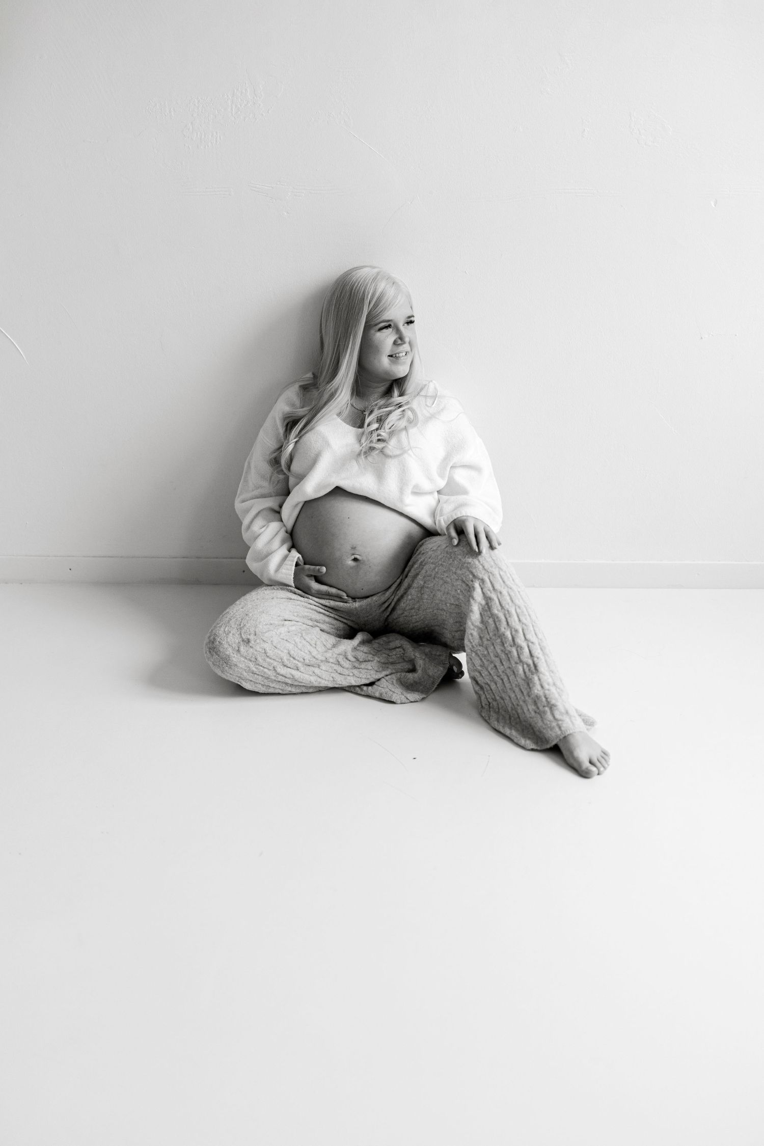 Black and white photo of a smiling pregnant woman sitting on floor, cradling her baby bump in cozy knit pants and crop sweater.