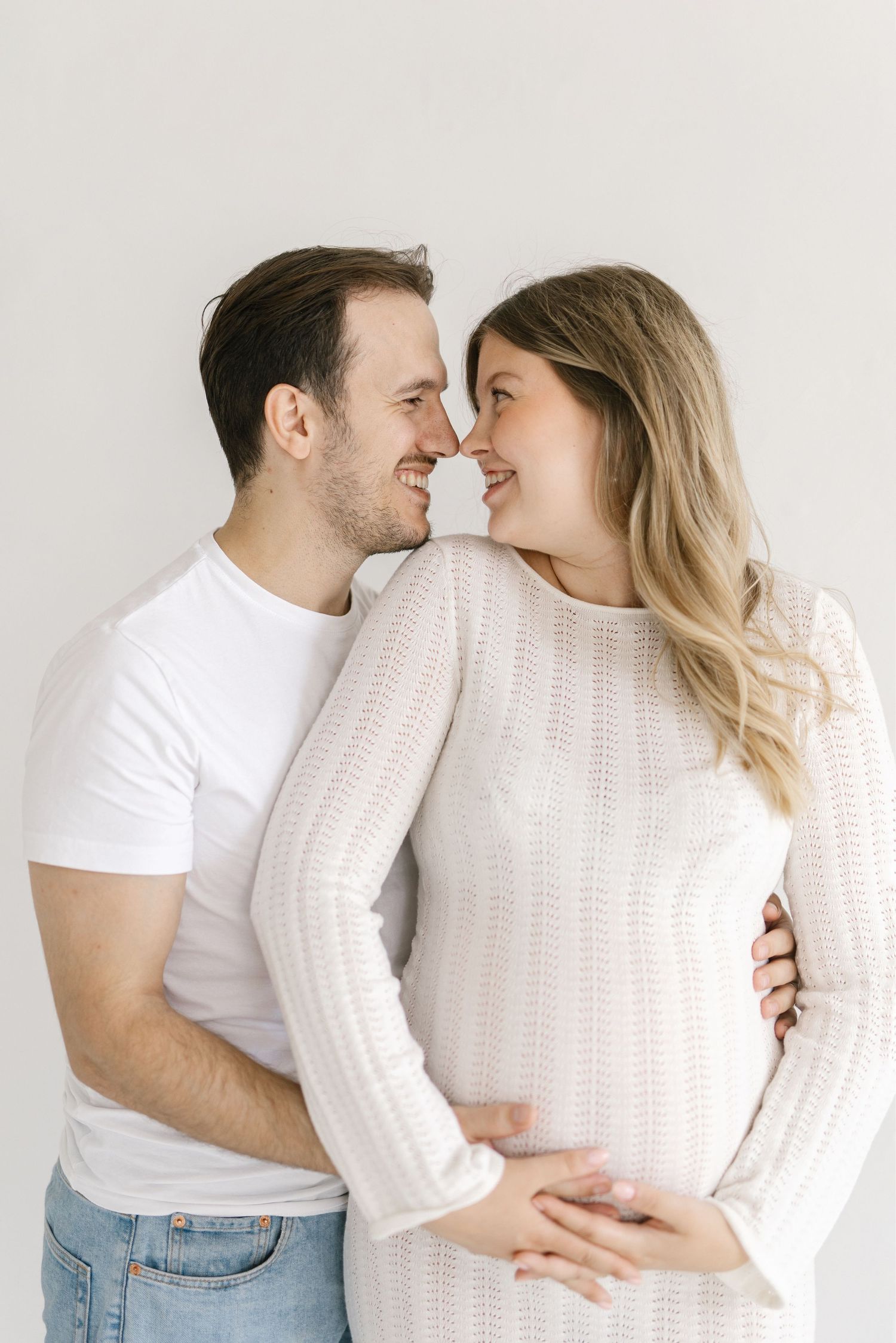 Happy couple sharing a tender moment, man embracing pregnant woman in white knit dress against light background.