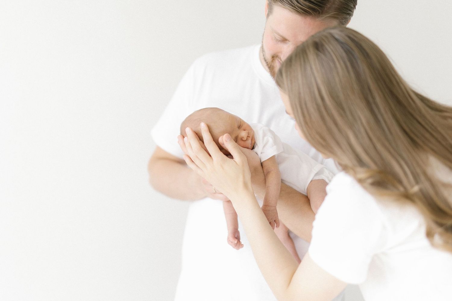 Two parents in white shirts tenderly holding their newborn baby against a soft white background.