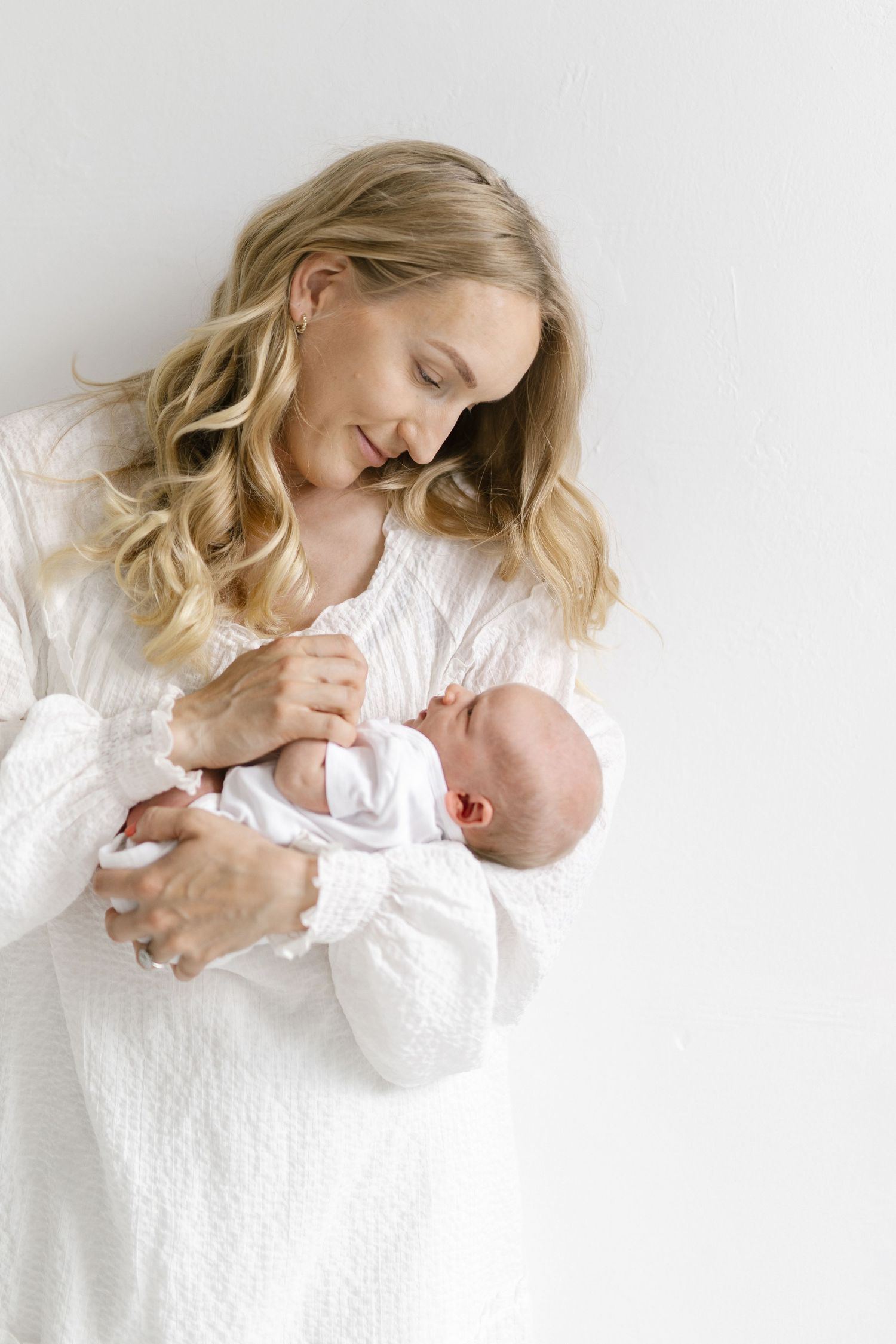 Parent in white lace dress holding and gazing lovingly at their newborn baby against light background.