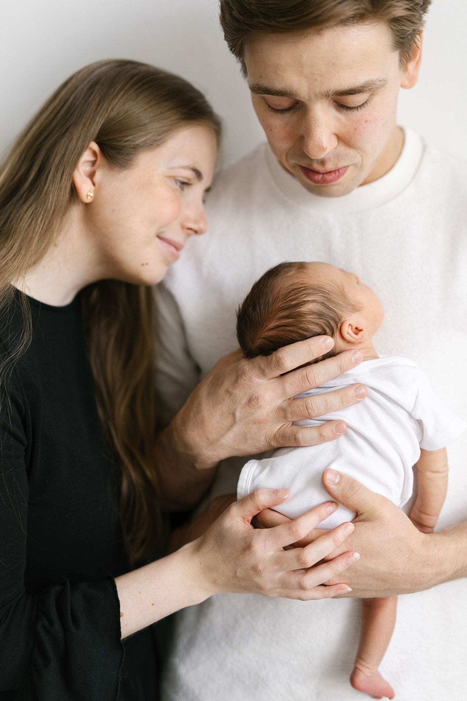 A young couple tenderly gazing at their newborn baby dressed in white, captured in a soft, neutral-toned portrait.
