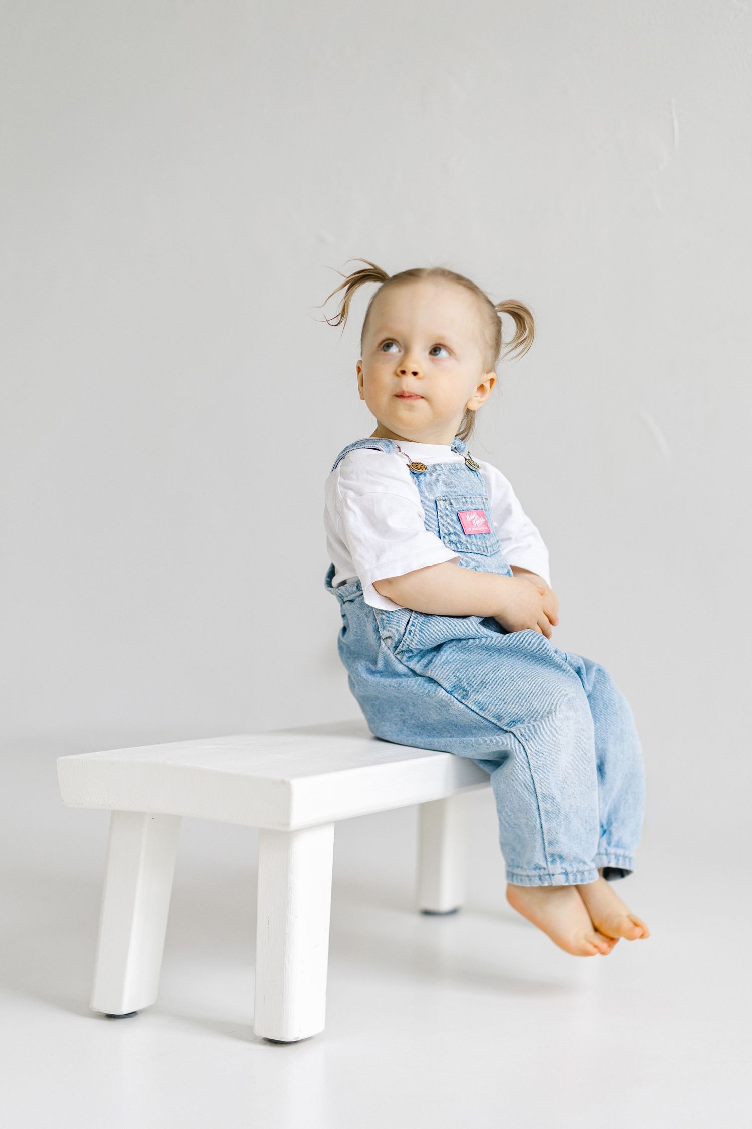 Cute toddler girl with pigtails sitting on a white bench, wearing denim overalls and a white shirt.