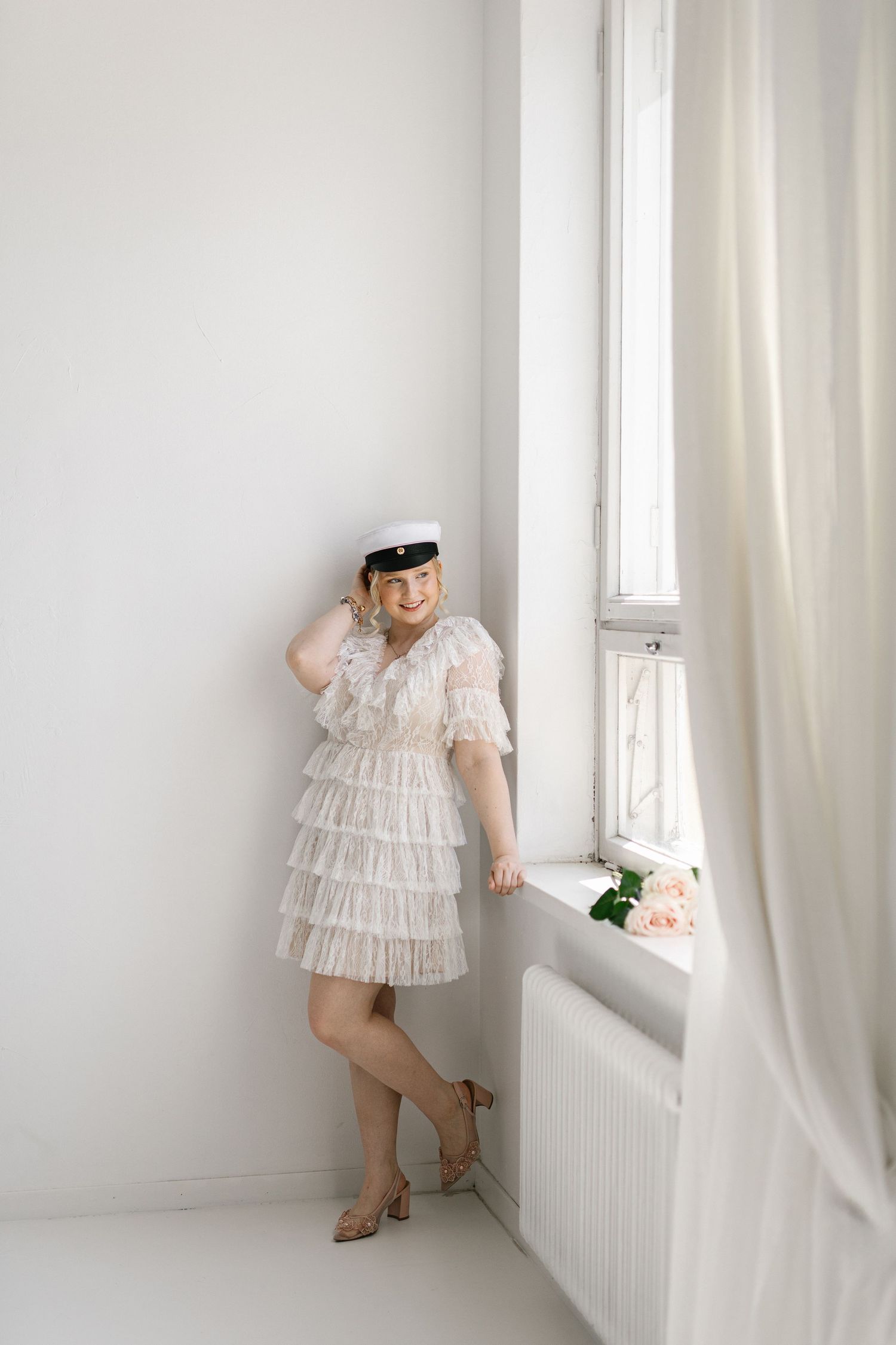 Woman in white ruffled dress and sailor cap poses by bright window with pink roses.