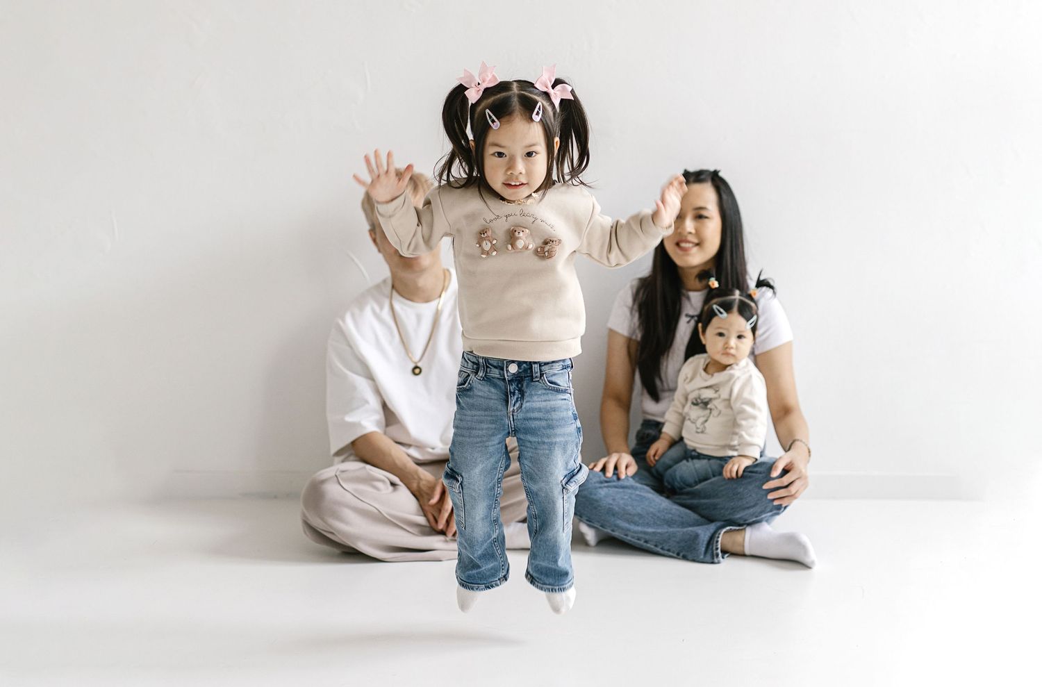 Asian family of four posing against white wall, toddler girl jumping with pigtails, parents seated with baby.
