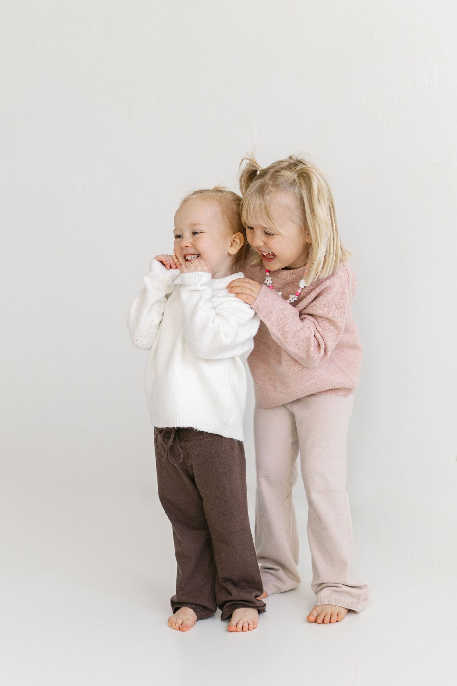 Two laughing toddlers in cozy neutral knitwear play barefoot against a white studio backdrop, one hugging the other from behind.