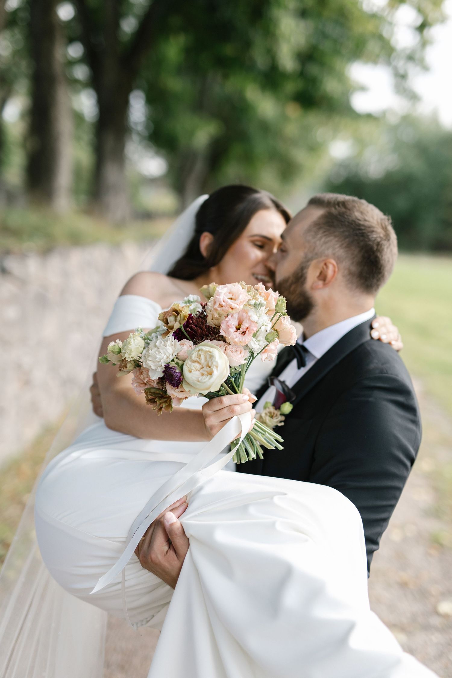 Groom carries bride in white off-shoulder gown holding floral bouquet, sharing a kiss outdoors near stone wall.