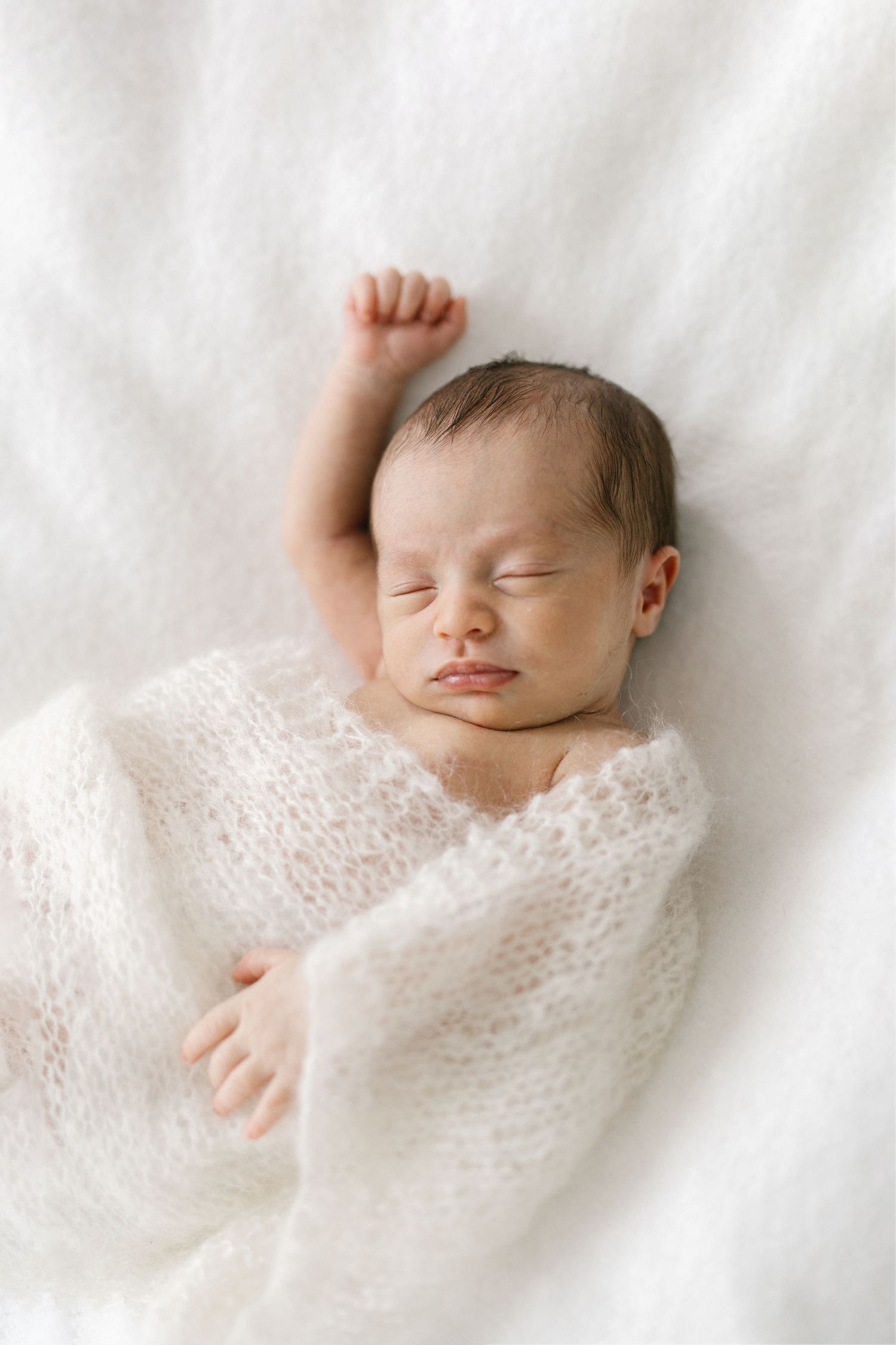 Peaceful newborn baby sleeping wrapped in a soft white knit blanket with one arm raised on a white fluffy background.