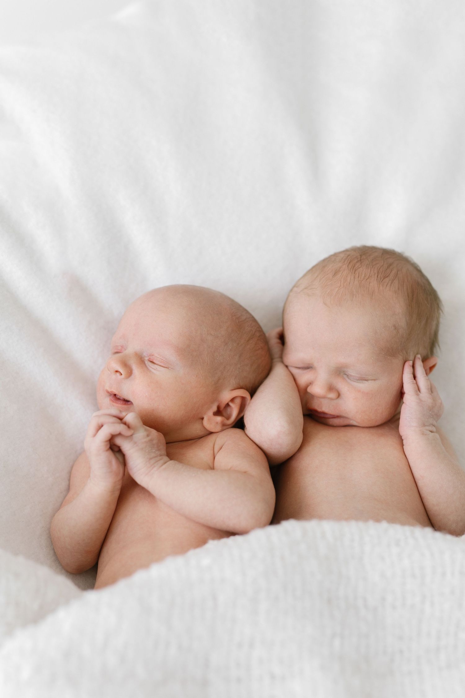 Two sleeping newborn twins lying side by side on a soft white blanket in a peaceful newborn photography pose.