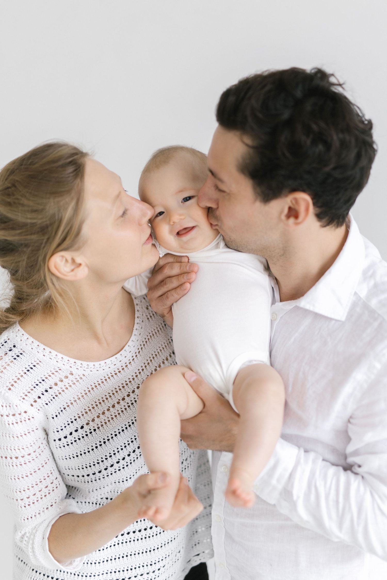 Happy parents kissing their smiling baby on the cheeks, dressed in white, against a light background.
