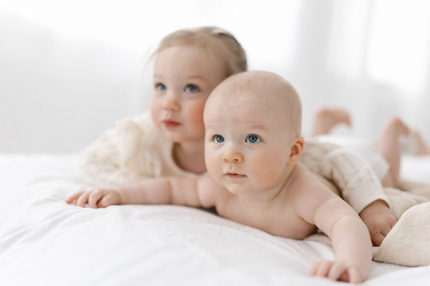 Two adorable babies with blue eyes lying on a white bed, one infant in a diaper and one toddler in a cream outfit.