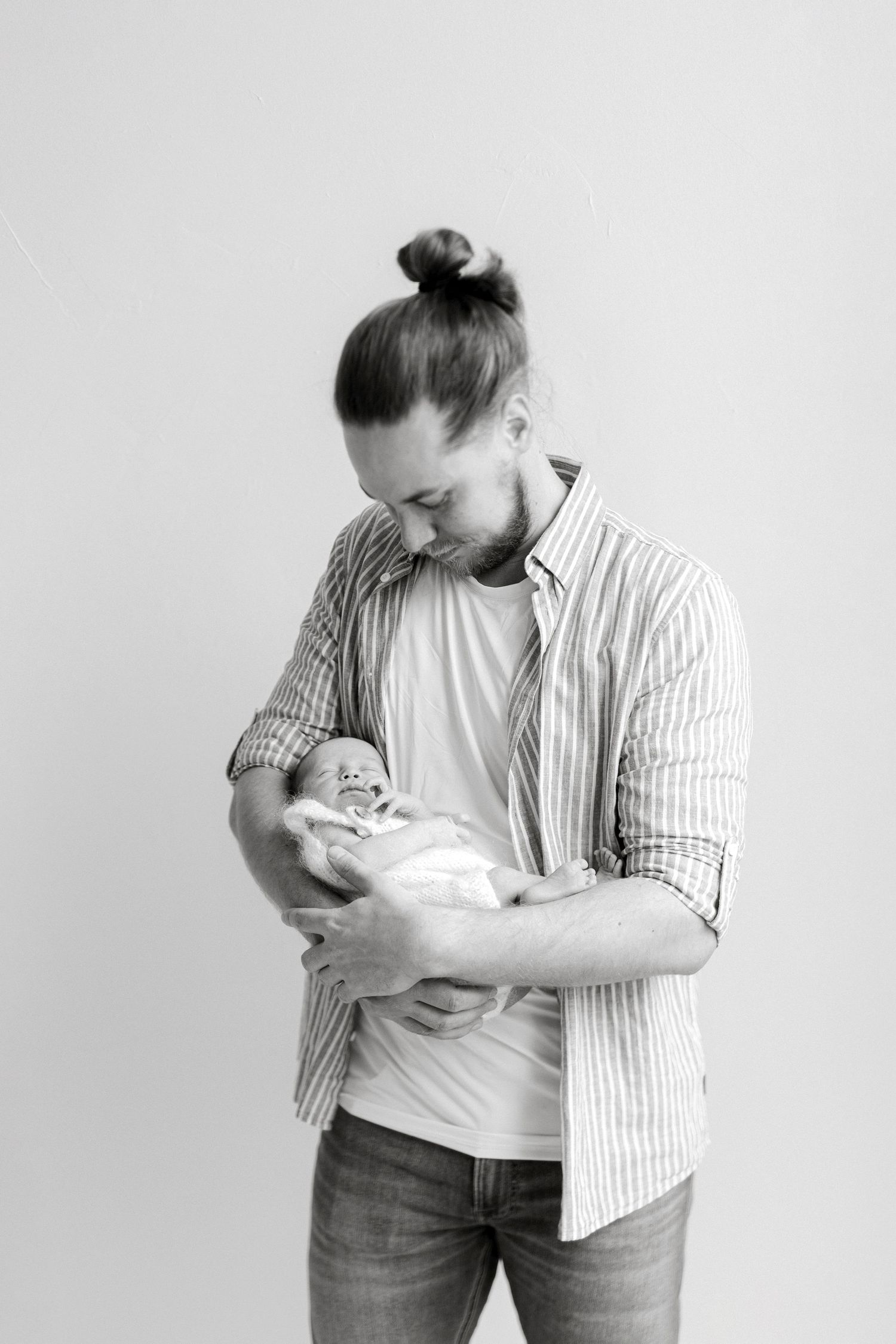 Black and white photo of a bearded man with a bun holding a newborn baby, looking down lovingly.