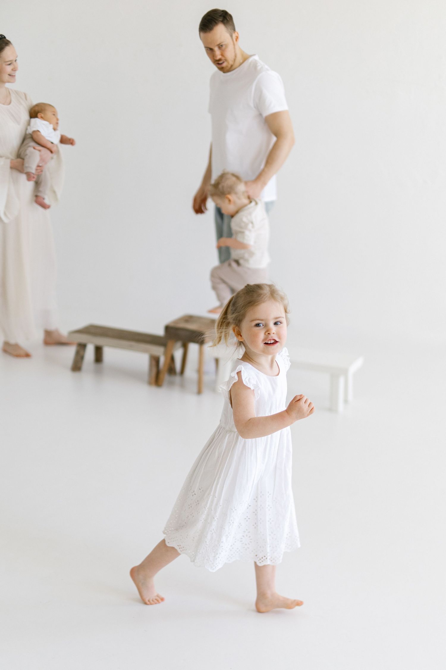 A joyful little girl in a white dress runs barefoot while her family poses together in a bright white studio.