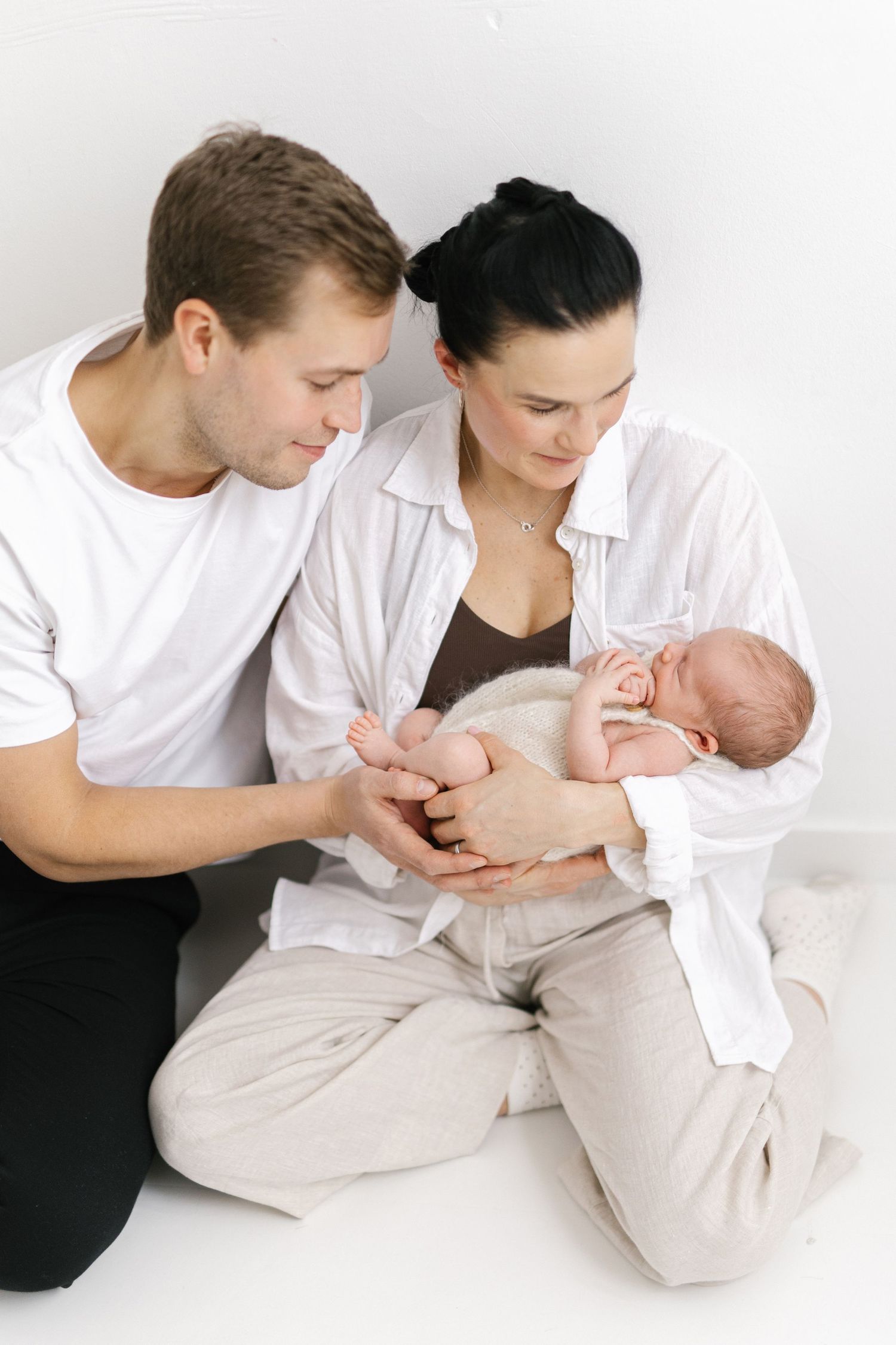 Young parents in white shirts sit on floor gazing lovingly at their newborn baby cradled in mother's arms.