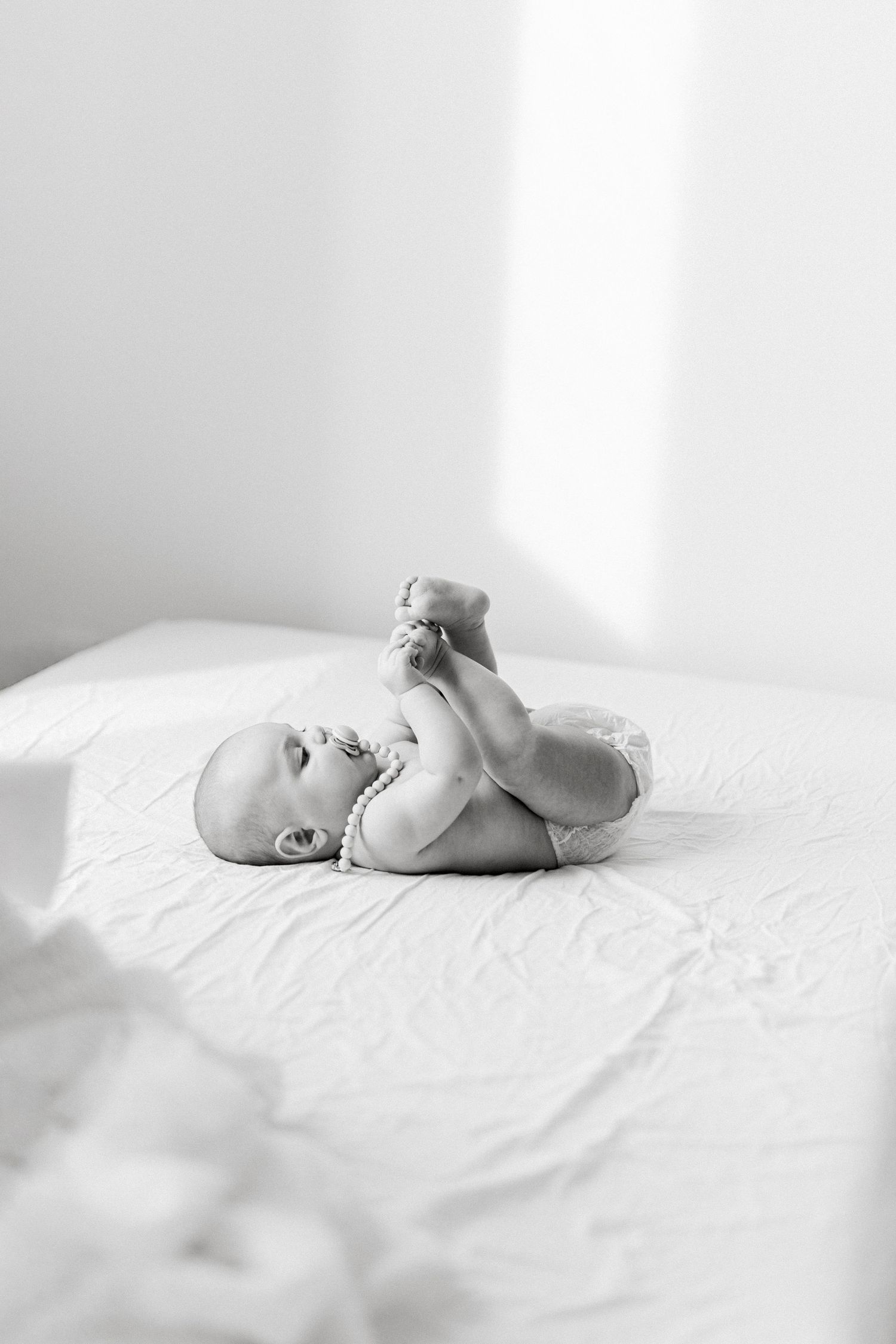 Black and white photo of a cute baby lying on white bed, holding feet up in the air.