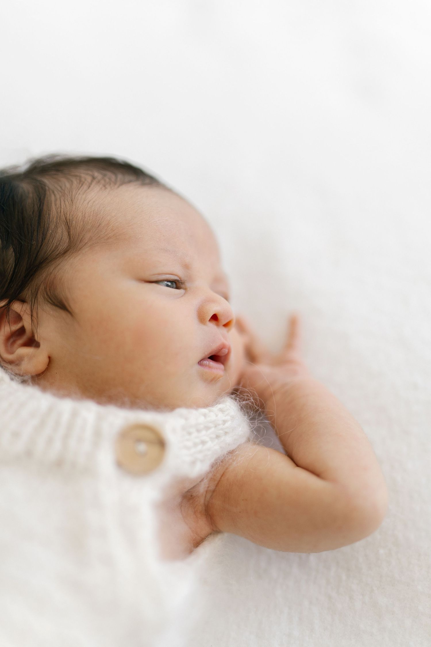 Newborn baby with dark hair lying on white blanket wearing a cream knit romper with wooden buttons.