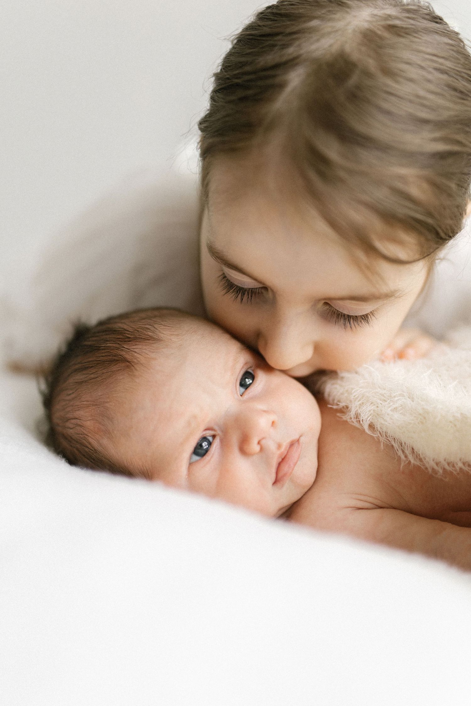 Older child gently kissing a newborn baby on the head while the infant looks up with bright eyes.