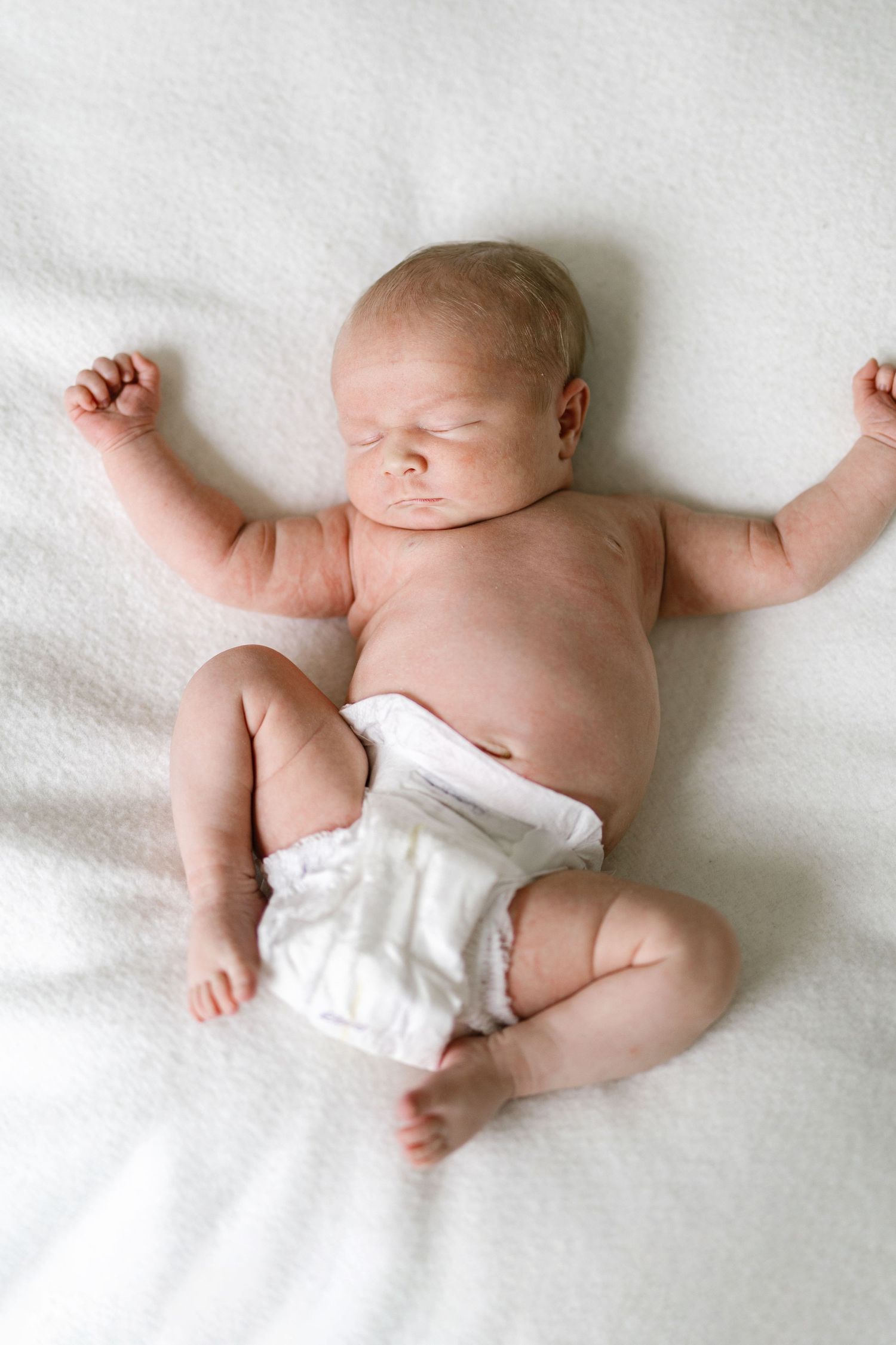 Newborn baby in white diaper lying on back on white blanket with arms raised, eyes closed, sleeping peacefully.