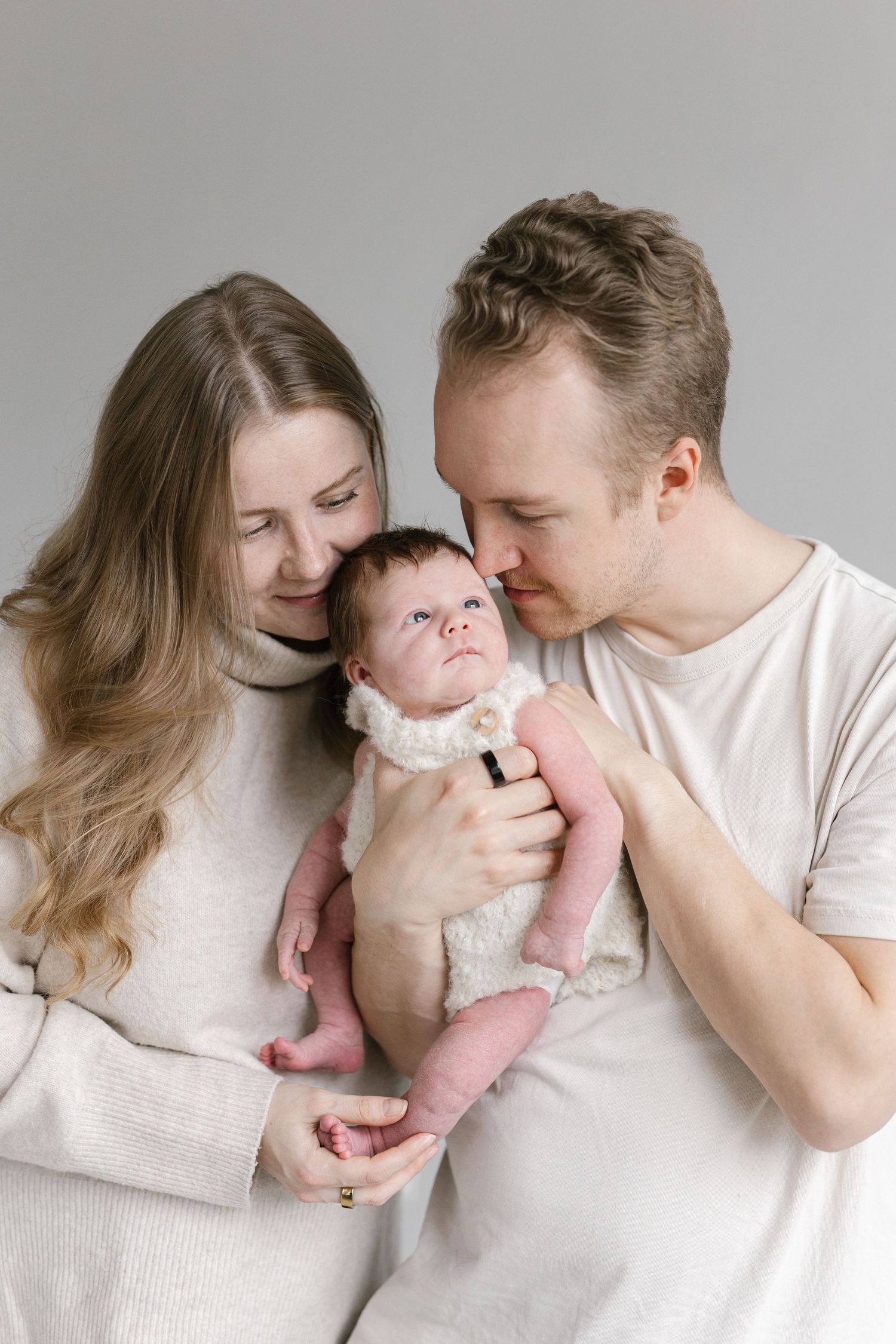 A smiling woman and a man gently gaze down at a newborn baby in a white knit outfit during a cozy indoor family portrait.