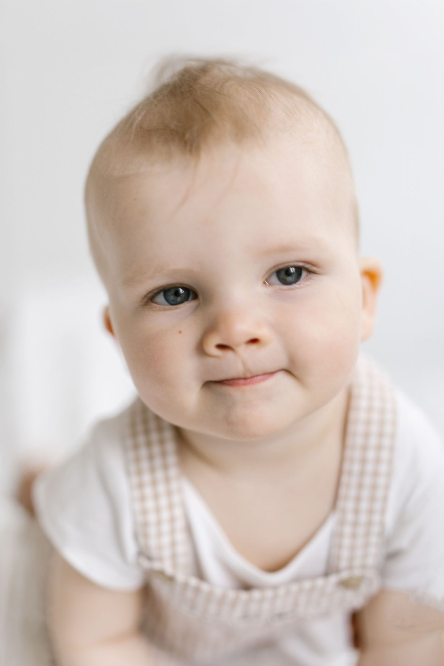 Adorable baby with blue eyes and light hair wearing a gingham overalls smiling softly against a white background.