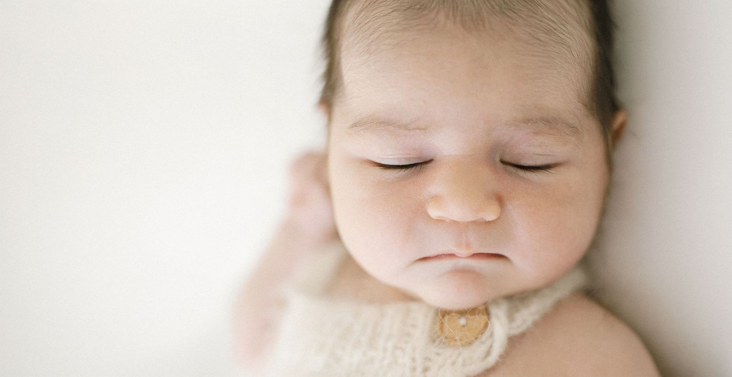 Sleeping newborn baby with a soft pout, wearing a cream knit romper with a wooden button against a light neutral background.