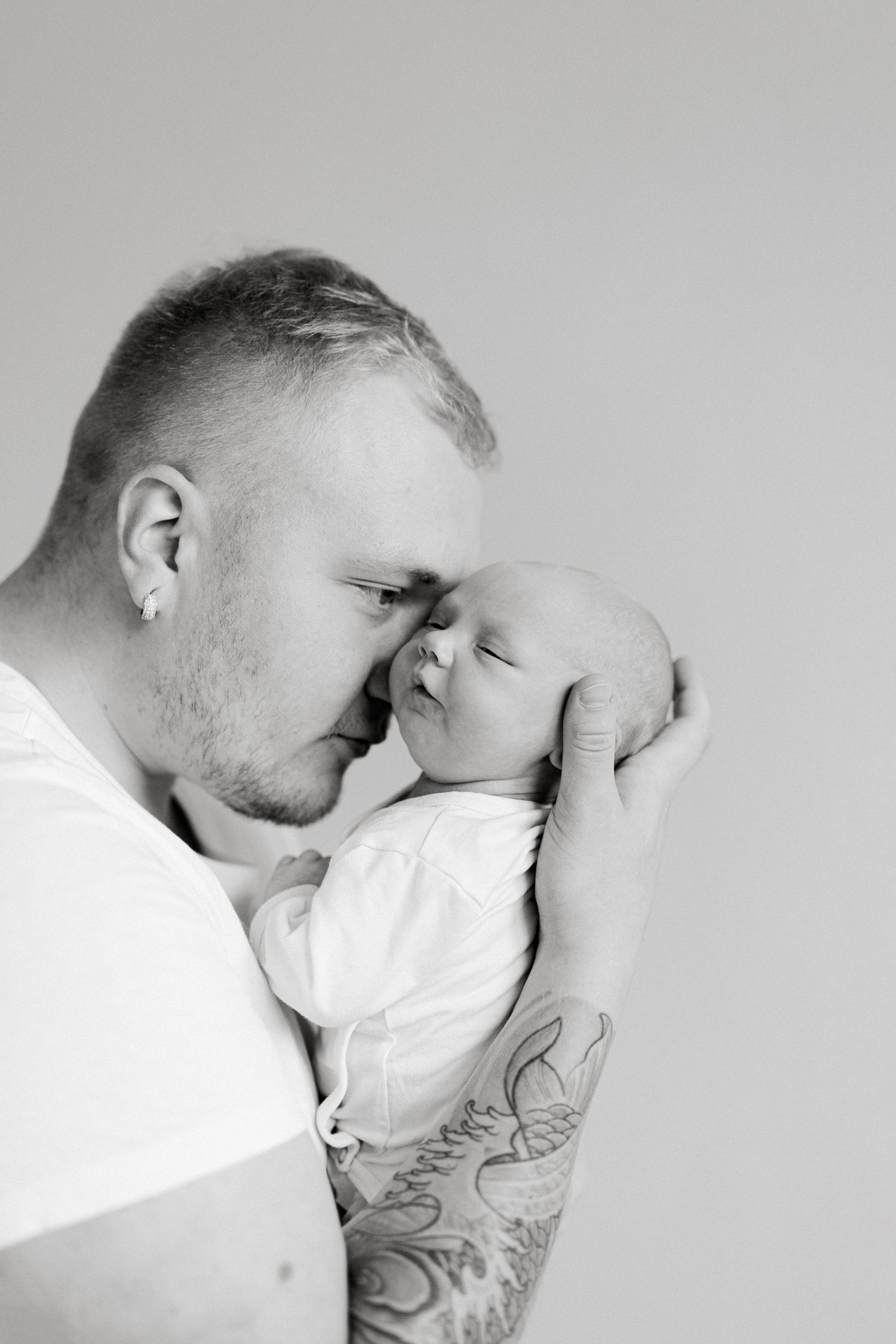 Black and white photo of a tattooed father tenderly kissing his sleeping newborn baby, held gently in his arms.