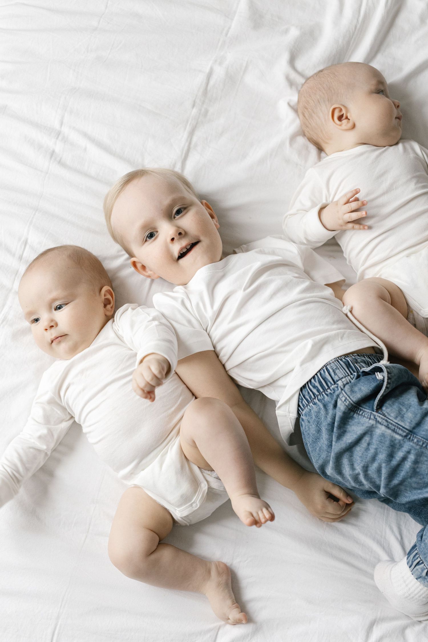 Three babies in white outfits lying together on white bedding.