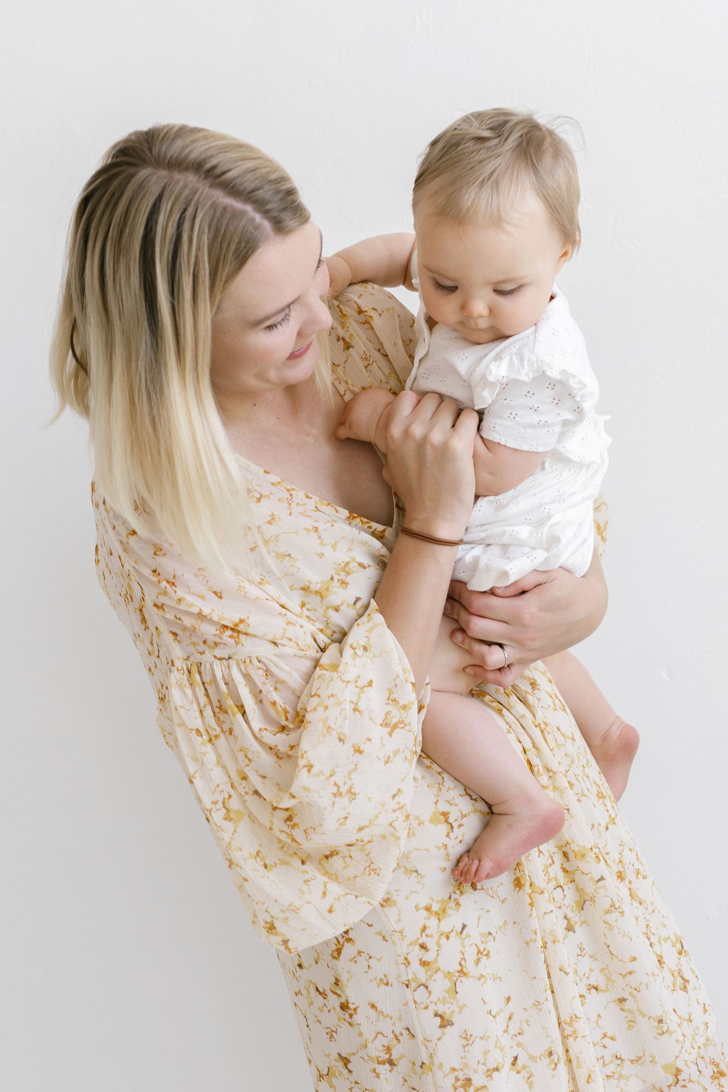 A smiling mother in a floral dress holds her baby in a white outfit against a light background.