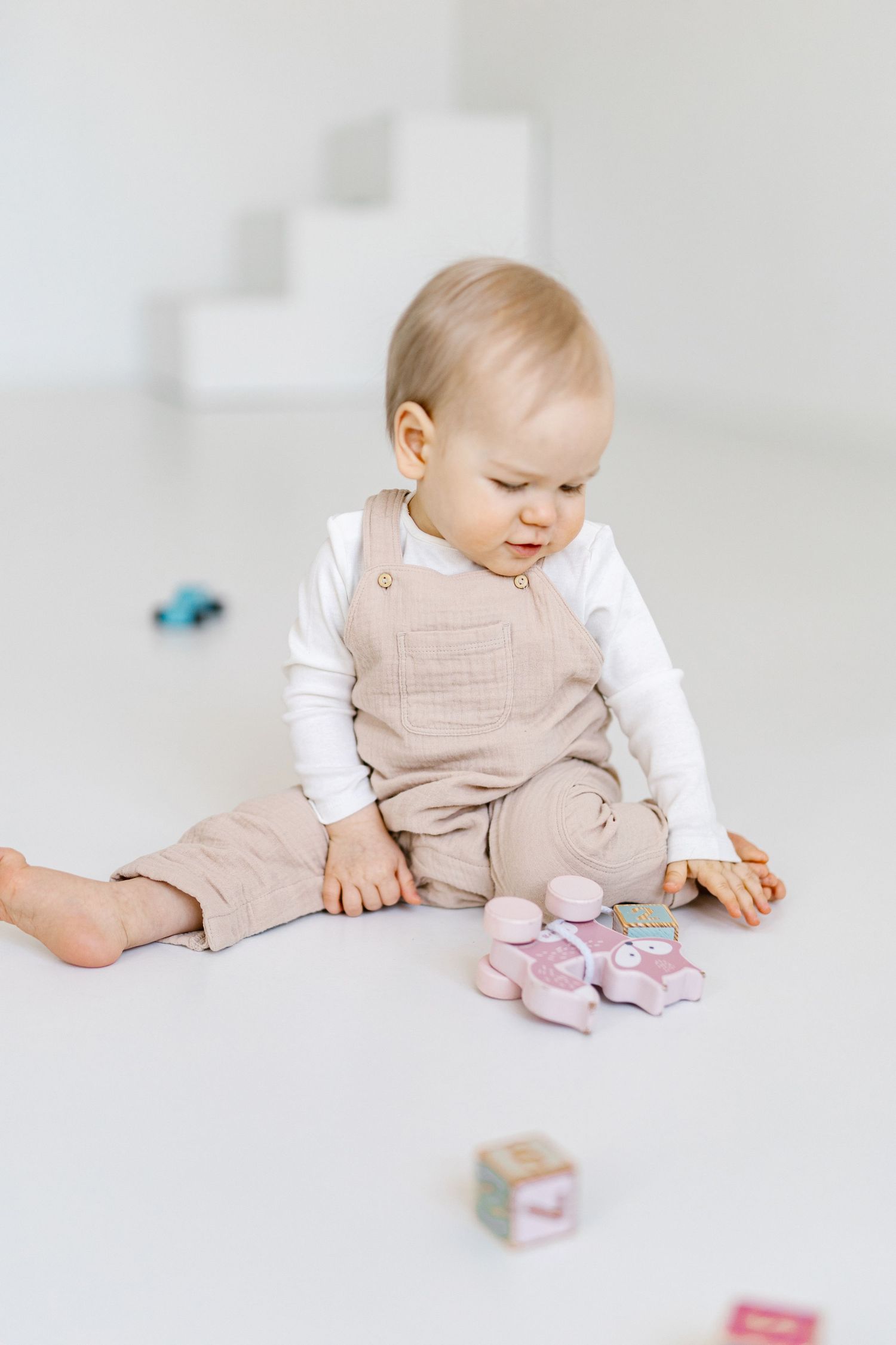 A baby in beige overalls sits on a white floor, playing with colorful toy blocks and small cars.