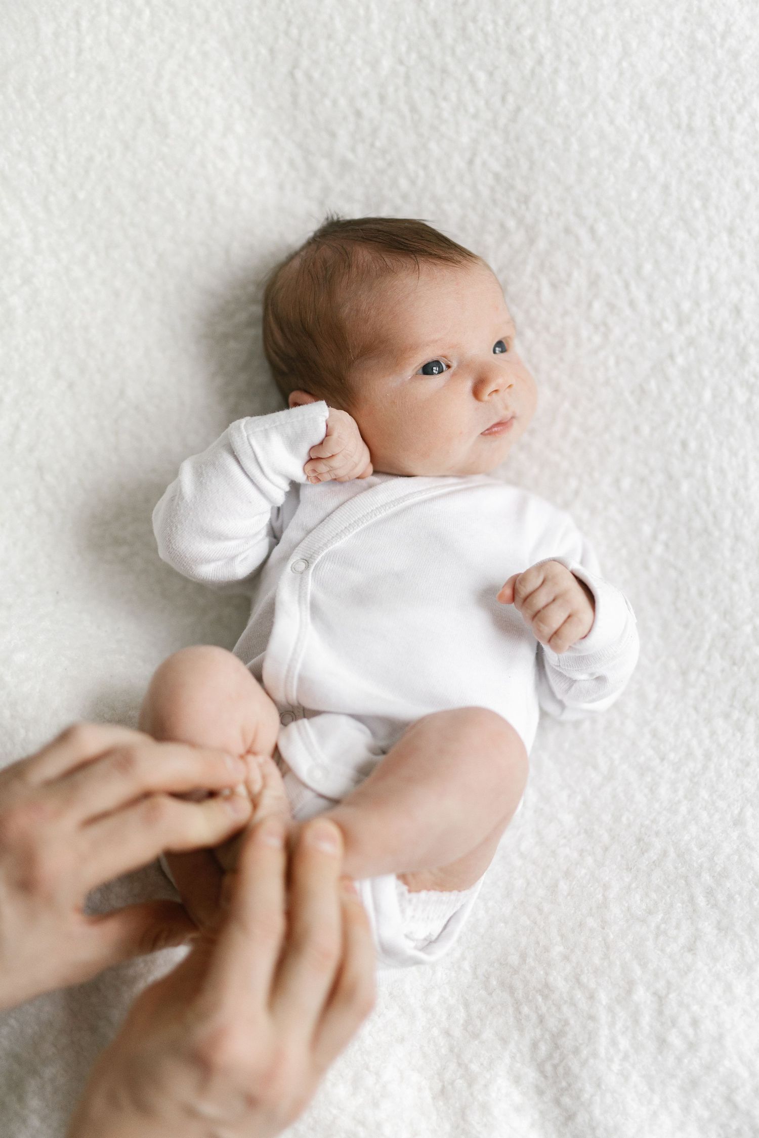 Baby in white onesie lying on textured white surface with hands reaching in.
