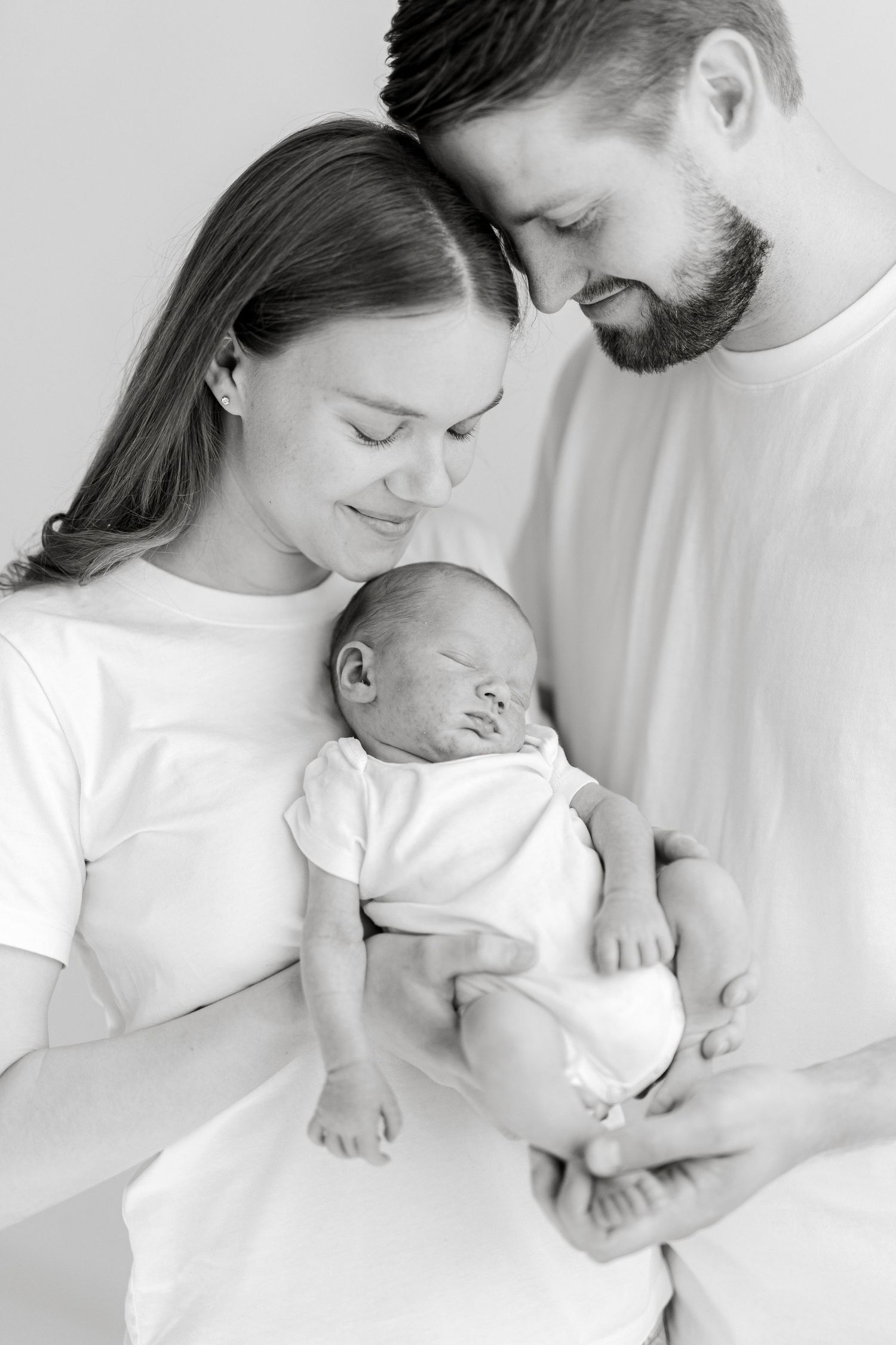 Black and white photo of new parents lovingly gazing at their sleeping newborn baby in a bright studio.