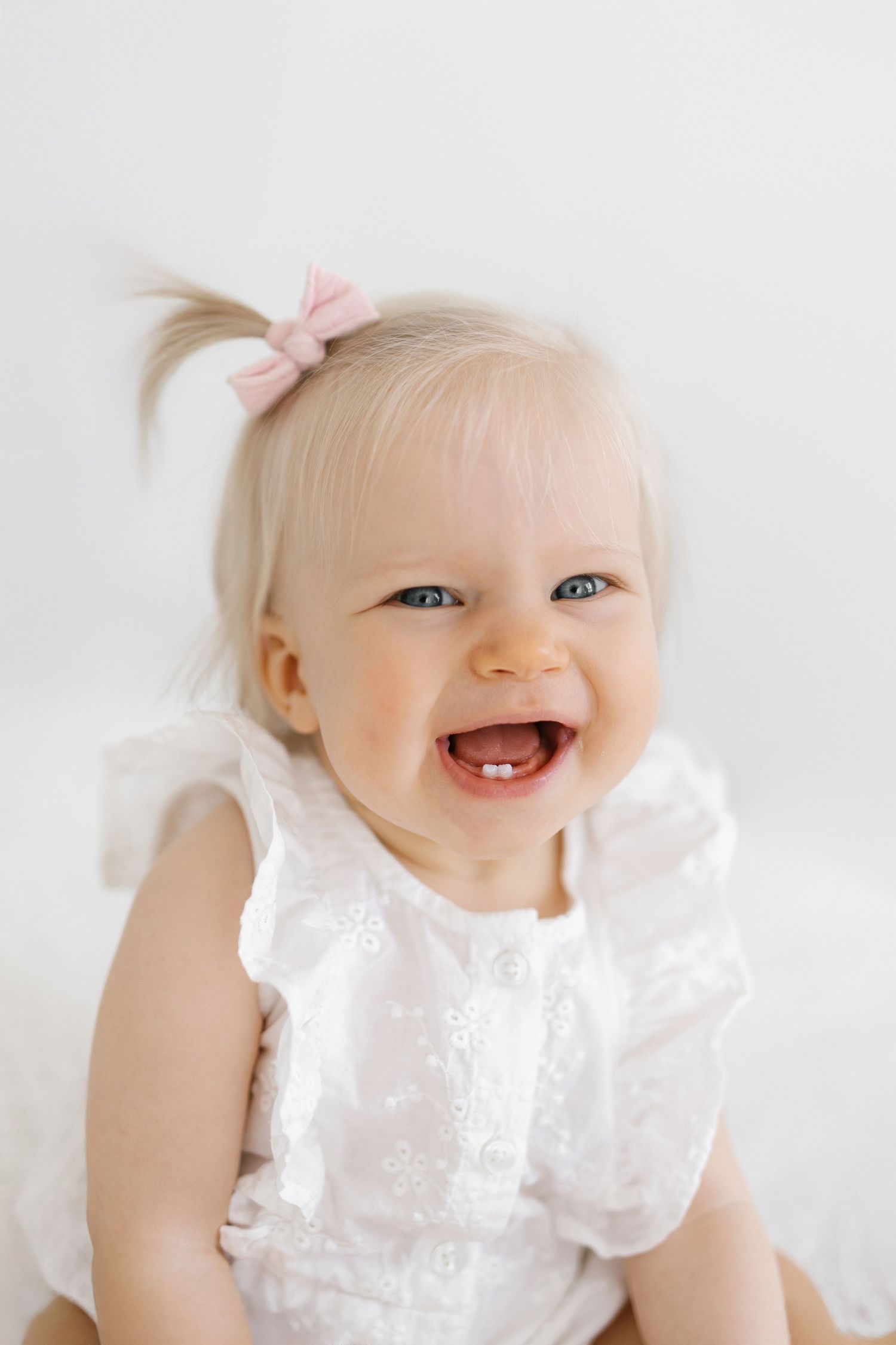 Baby in white lace dress with pink bow smiling and laughing.