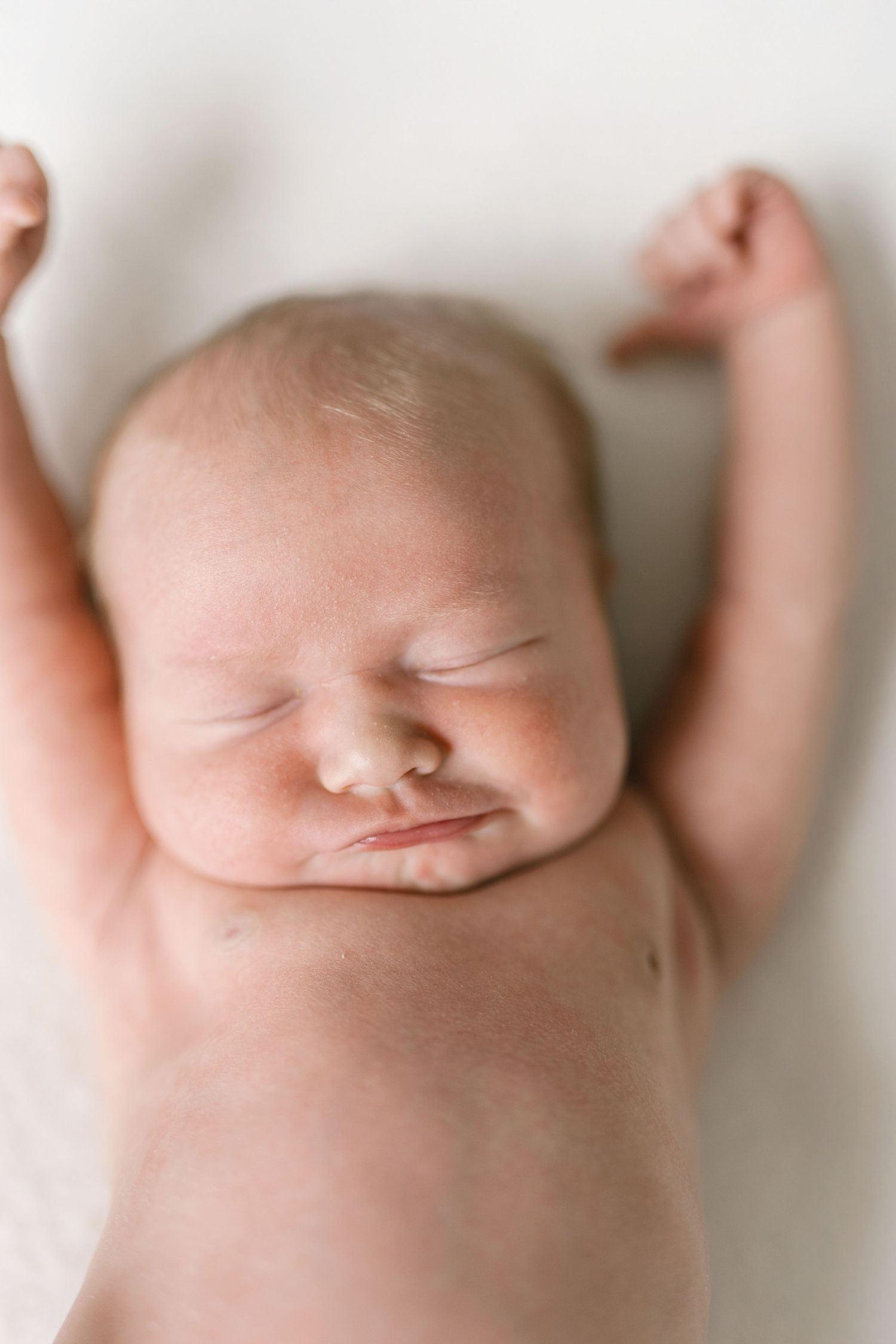 Peaceful sleeping newborn baby with arms raised overhead lying on white soft blanket in natural light.