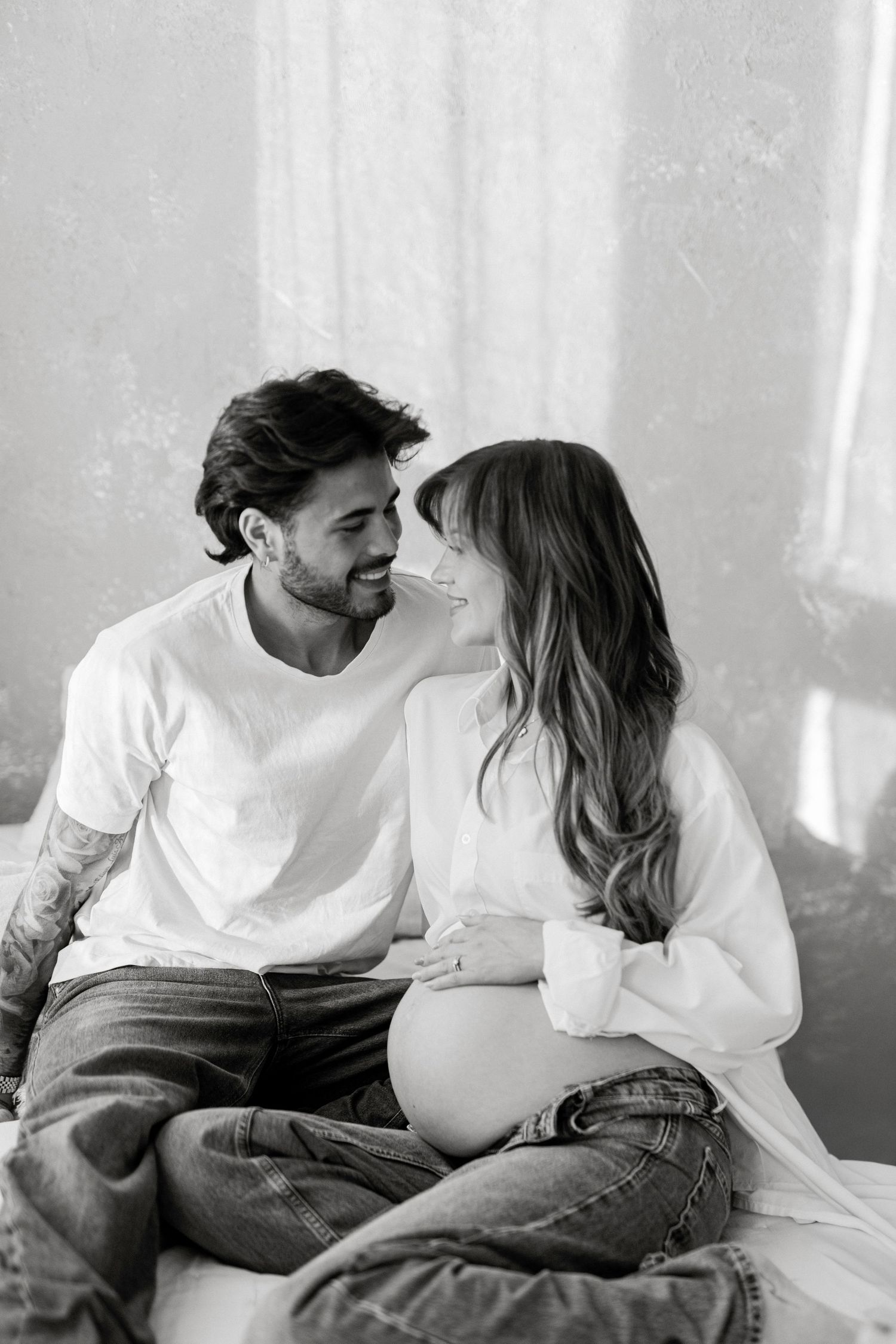 Black and white photo of a smiling couple sitting together, woman showing pregnant belly, man with tattoos gazing lovingly.