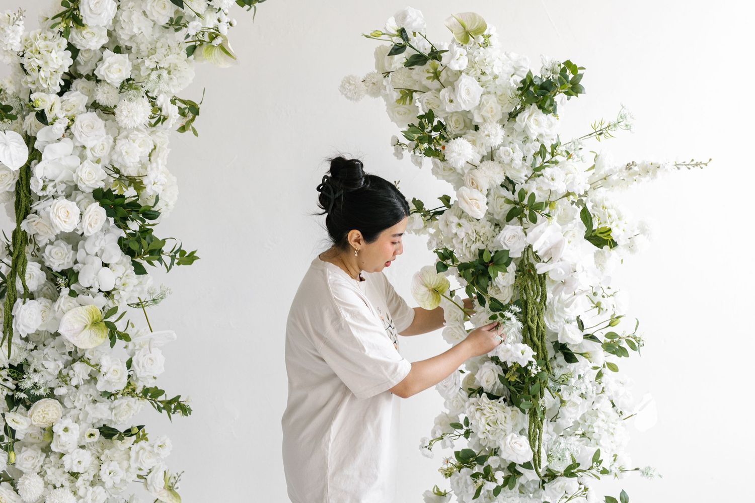 Woman in white outfit arranging white floral arch with roses and greenery for elegant wedding decor.
