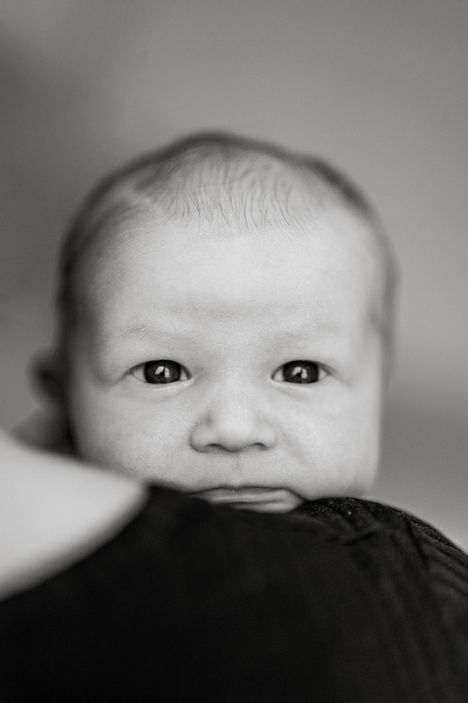 Black and white close-up portrait of a newborn baby with wide open eyes looking directly at the camera.