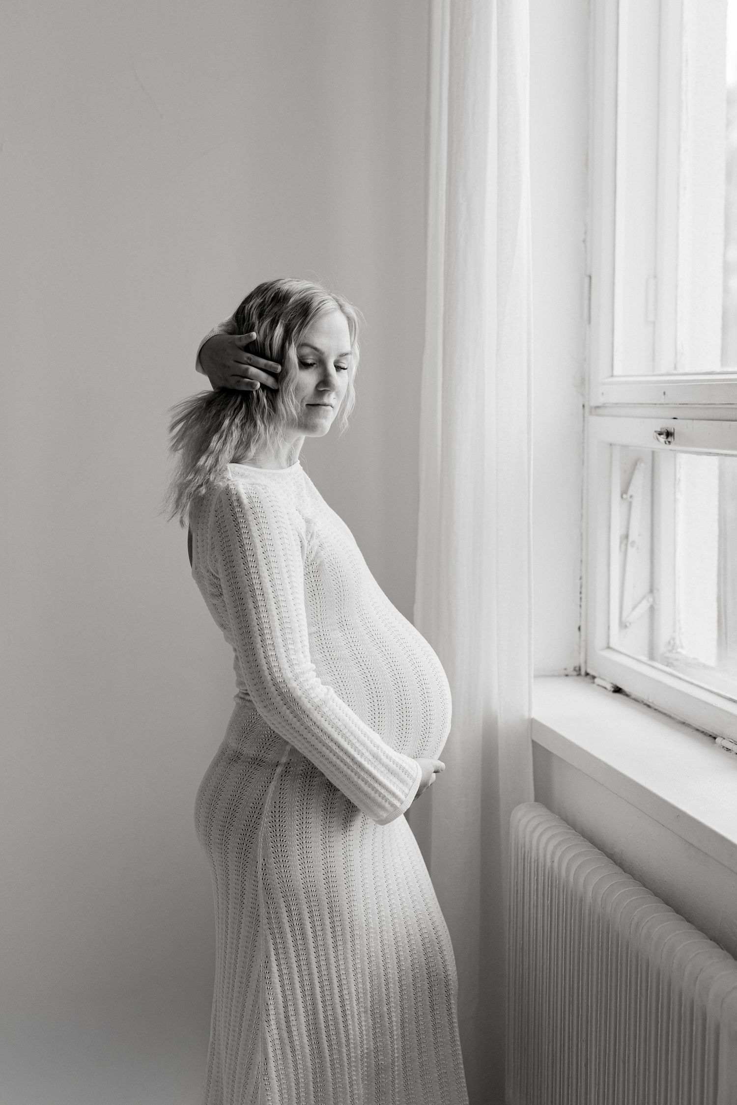 Black and white photo of a pregnant woman in a ribbed dress cradling her belly by a window.