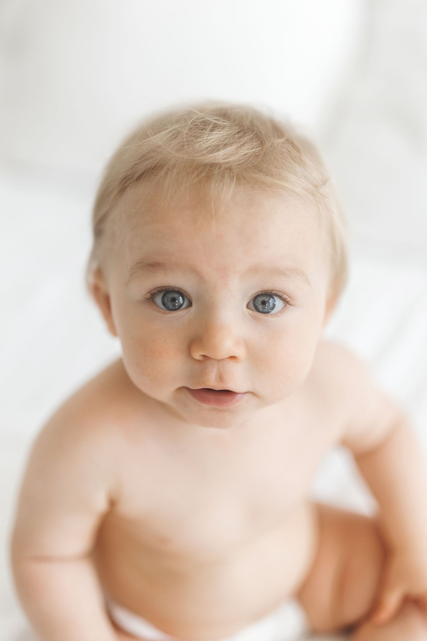 Adorable blue-eyed baby with blonde hair sitting up, looking curiously at camera against white background.