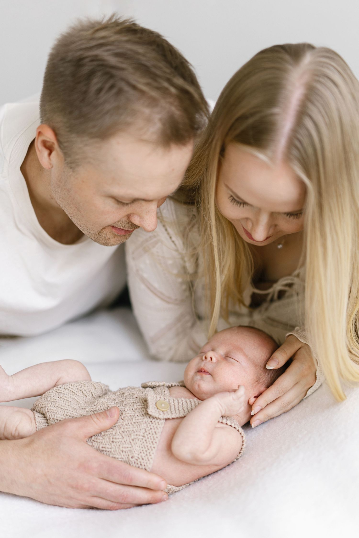 Parents lovingly gazing at their sleeping newborn baby dressed in a beige knit outfit on a white background.