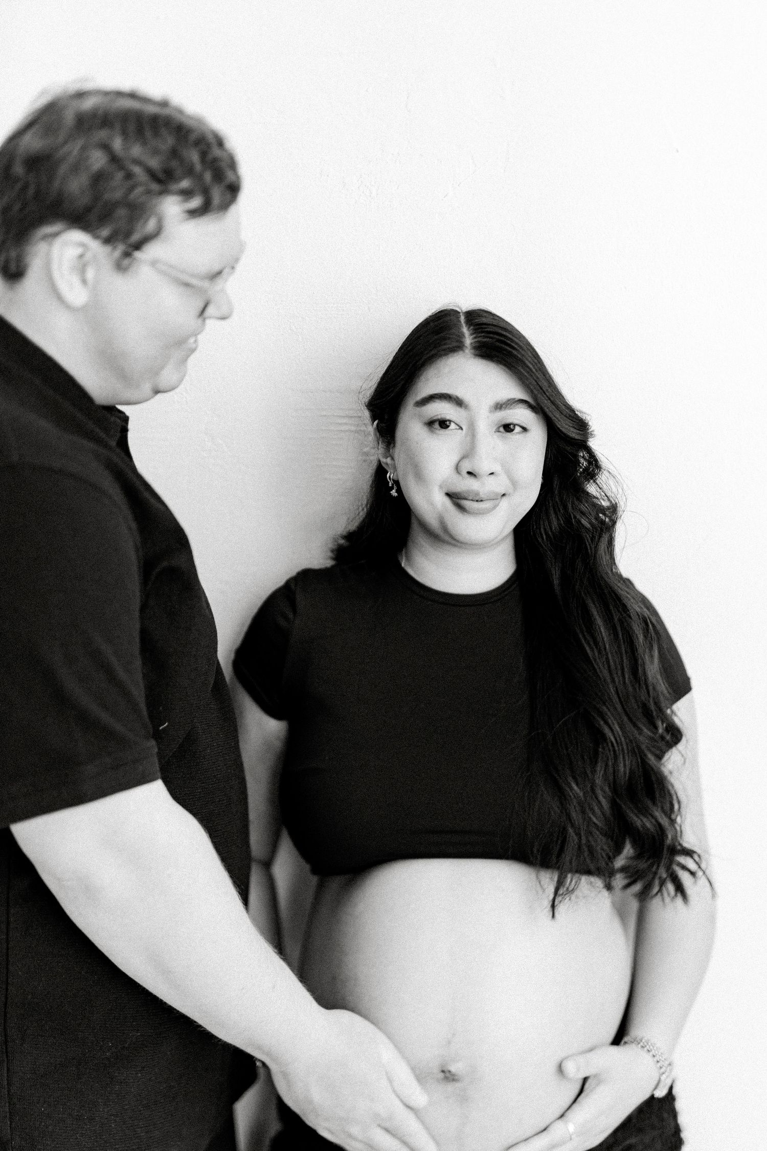 Black and white maternity portrait of an expectant couple, with a smiling woman in a crop top and her partner gently cradling her baby bump.