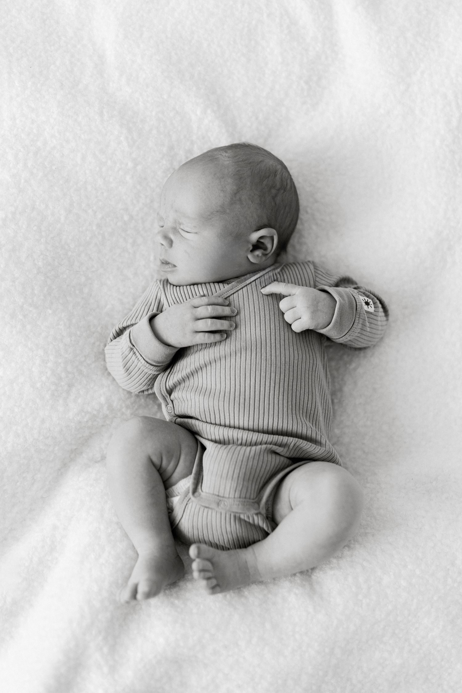 Black and white photo of a sleeping newborn baby wearing a ribbed onesie on white bedding.