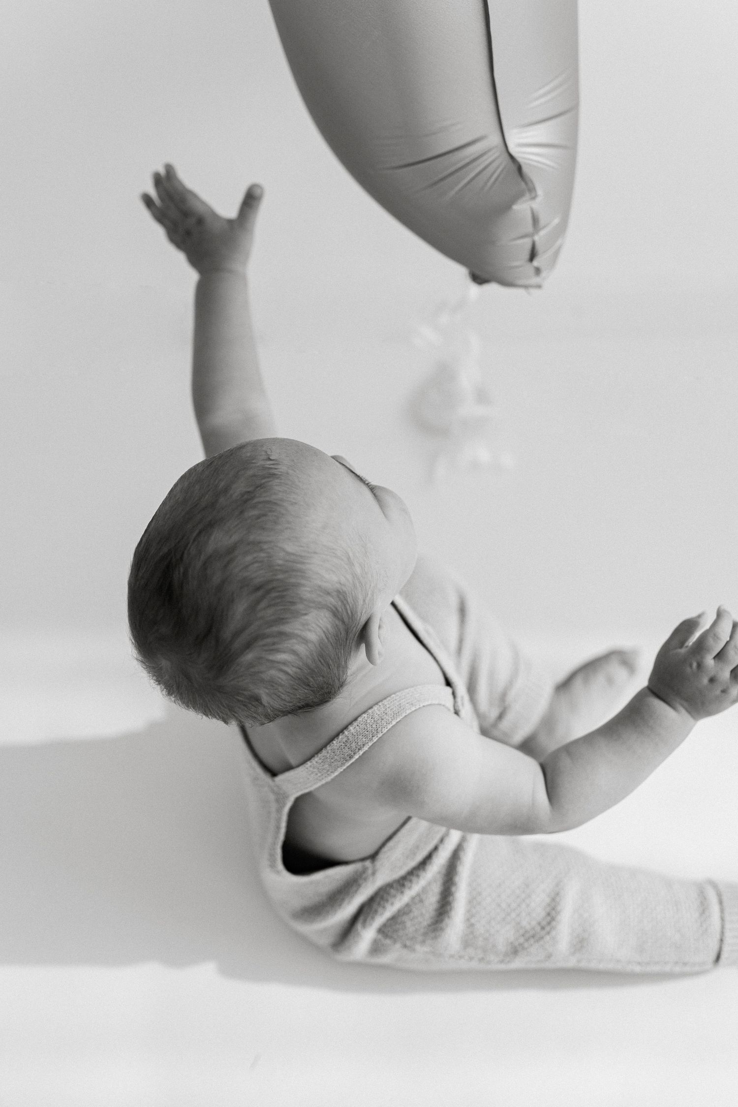 Baby reaching up towards floating balloon in artistic black and white photo.